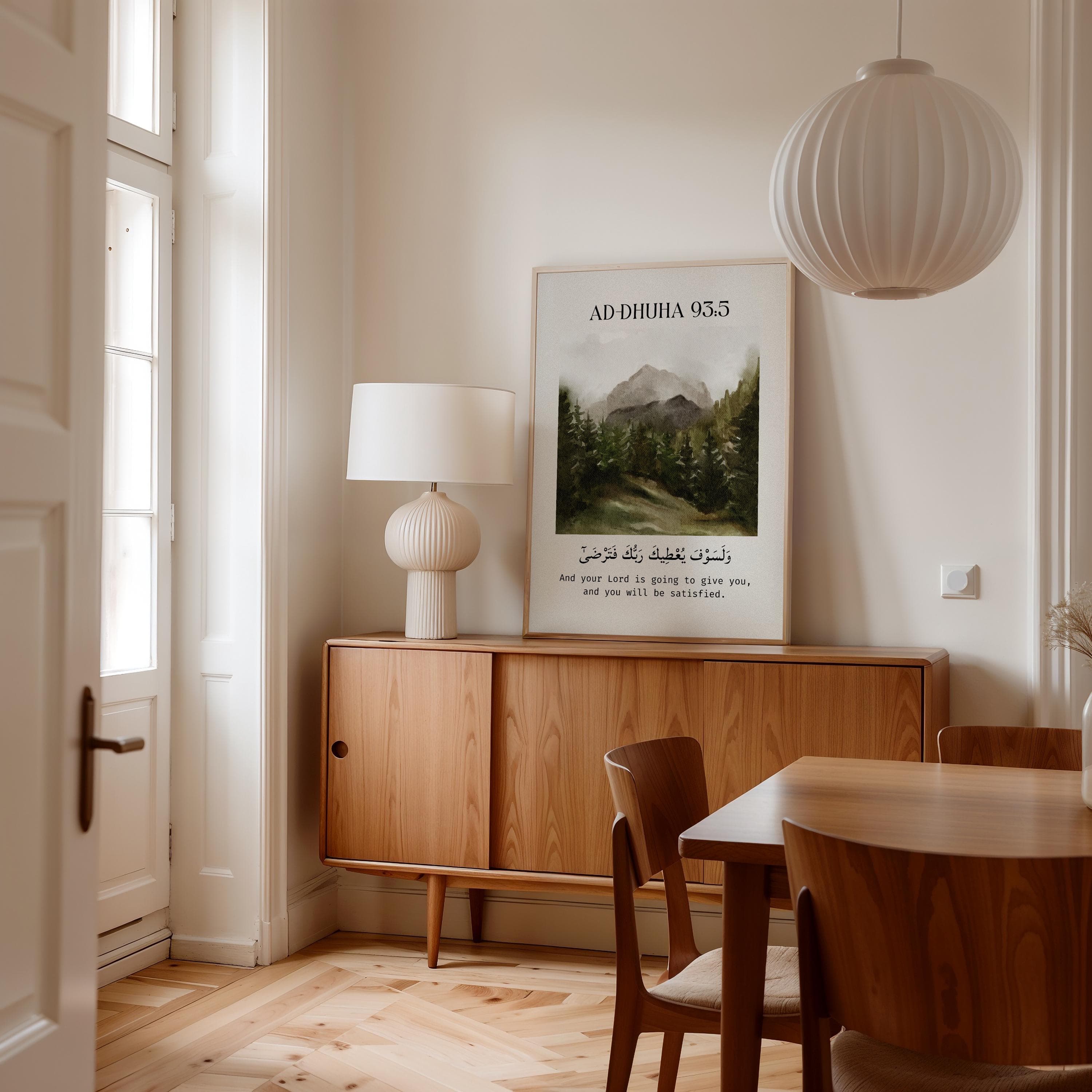 Islamic wall art print featuring Surah Ad-Dhuha 93:5 with Arabic and English translation: “And your Lord is going to give you, and you will be satisfied.” Centered above a watercolor-style landscape of mountains and pine trees, framed in light wood against a minimalist off-white wall, capturing a serene and spiritual vibe.
