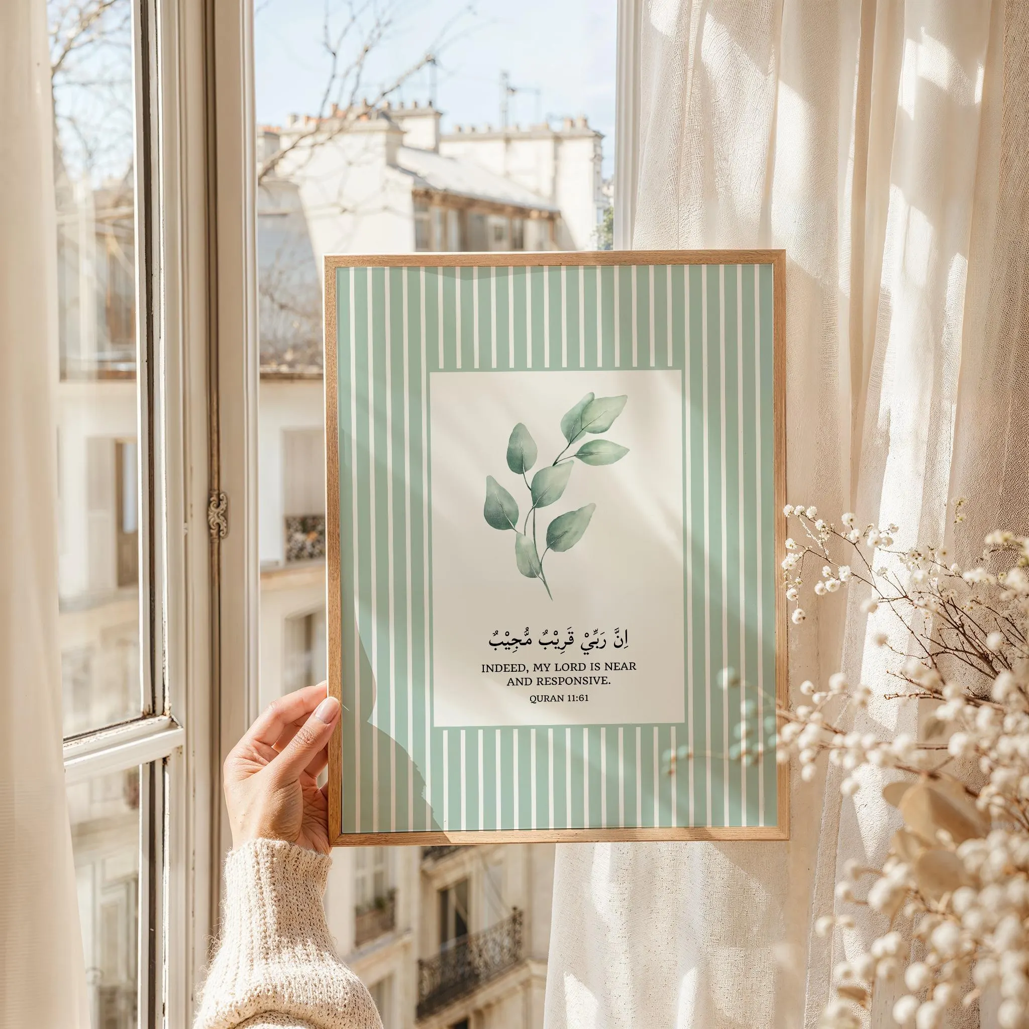 Vertical botanical illustration of a leafy green eucalyptus branch in soft watercolors. The central white art panel is surrounded by a sage green and white vertical pinstripe border. Below the leaves, black Arabic calligraphy reads "Inna rabbi qaribun mujib," with the English translation: "Indeed, my Lord is near and responsive. Quran 11:61."