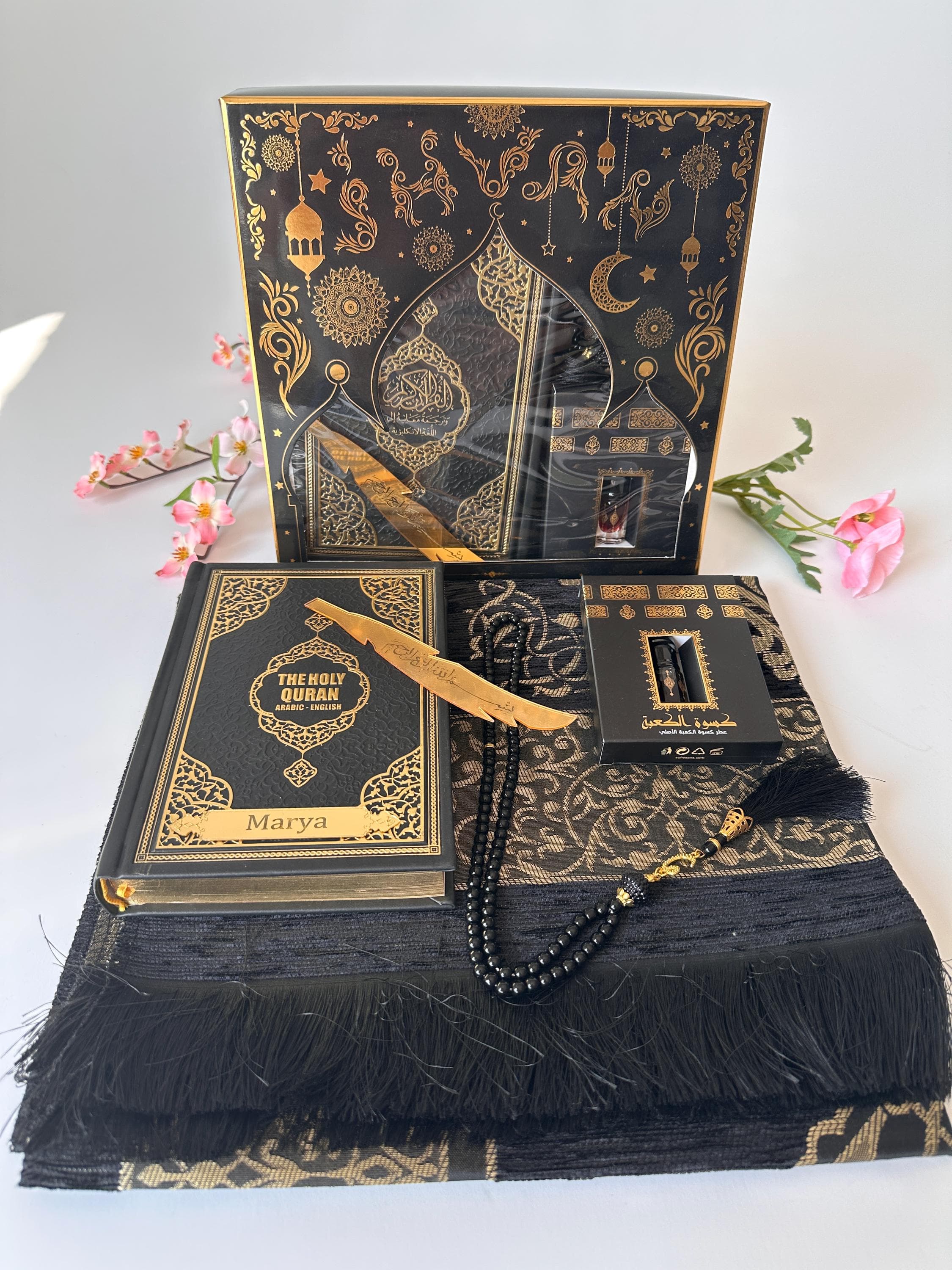 May include: Black and gold Islamic prayer set.  The set includes a prayer mat with gold detailing, a black Holy Quran with gold lettering and the name Marya, a prayer bead mala, a gold-colored bookmark, and a small black case containing a pen. The packaging is a black box with gold Islamic-inspired designs. The set is presented on a white background.