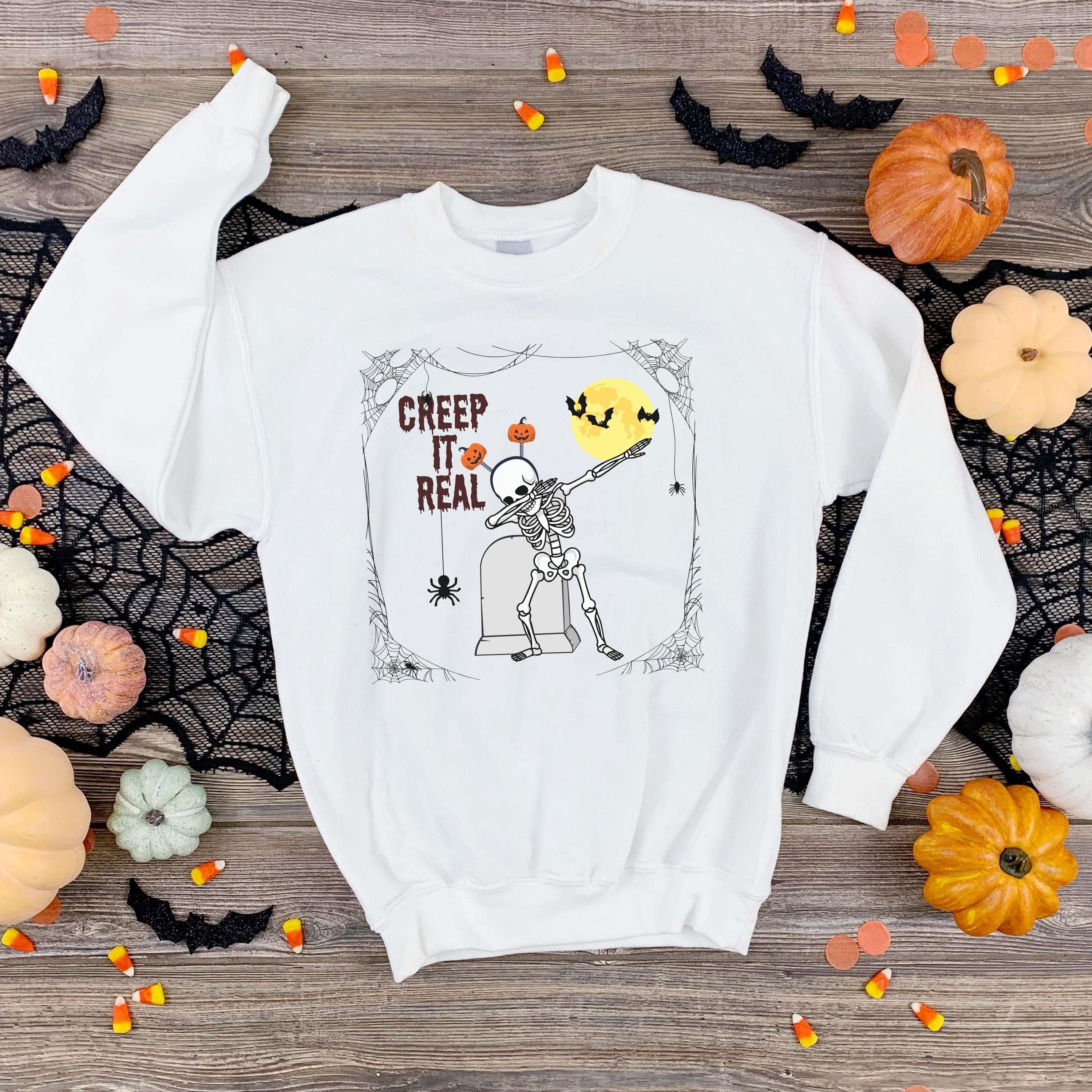 CREEP IT REAL funny halloween PNG Download, showing skeleton in a pose infront of a tombstone, moon with bats, spider and cobweb.