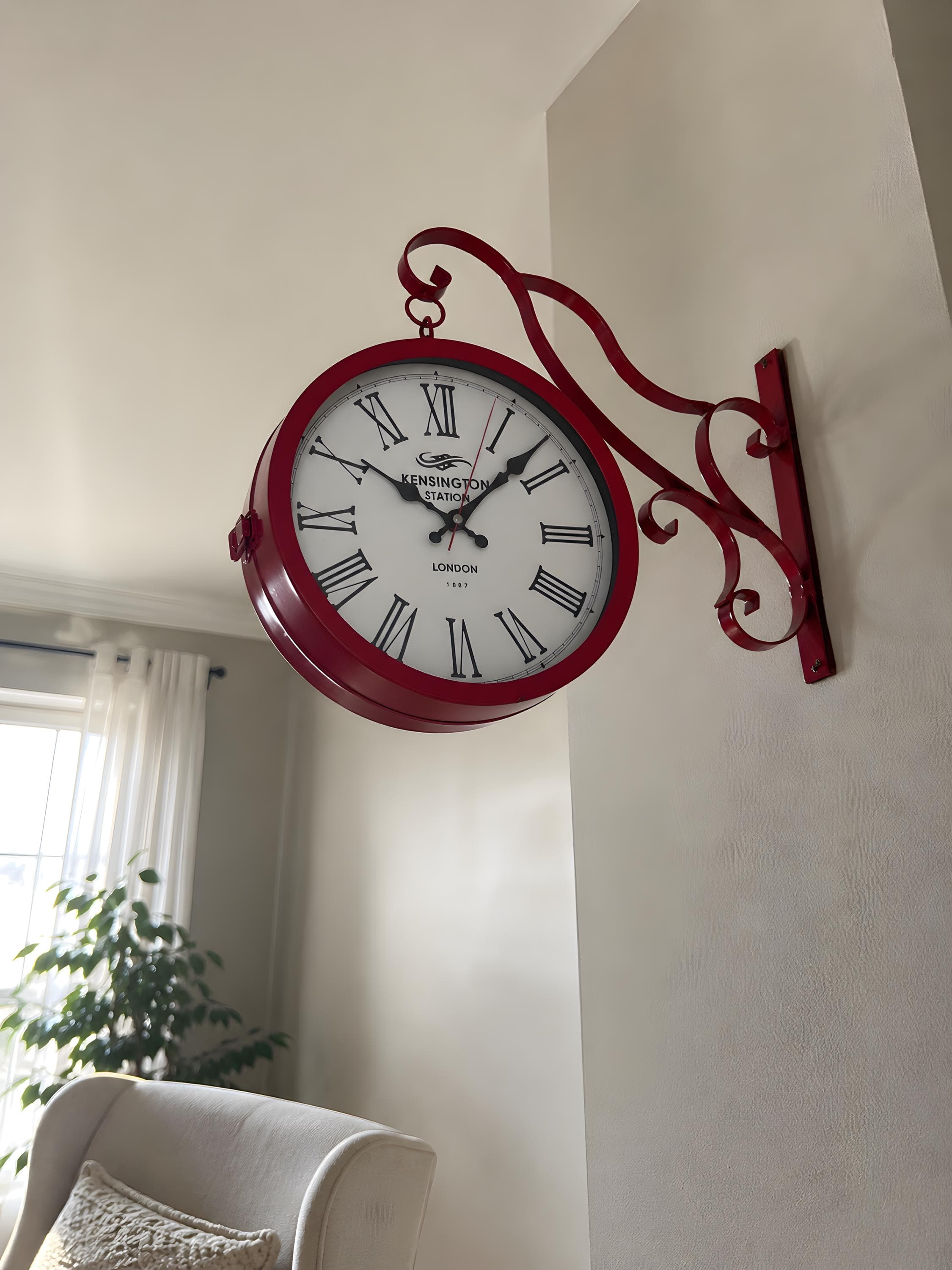 Crimson red Victorian railway station wall clock with ornate iron 
bracket installed on beige kitchen wall near open wooden shelving. 
Kensington Station London 1887 clock with 30 cm white Roman 
numeral dial and red second hand. Classic double-sided antique 
station clock as kitchen or hallway decor statement. Decorative 
scrollwork bracket adds vintage European railway character to 
everyday residential spaces.