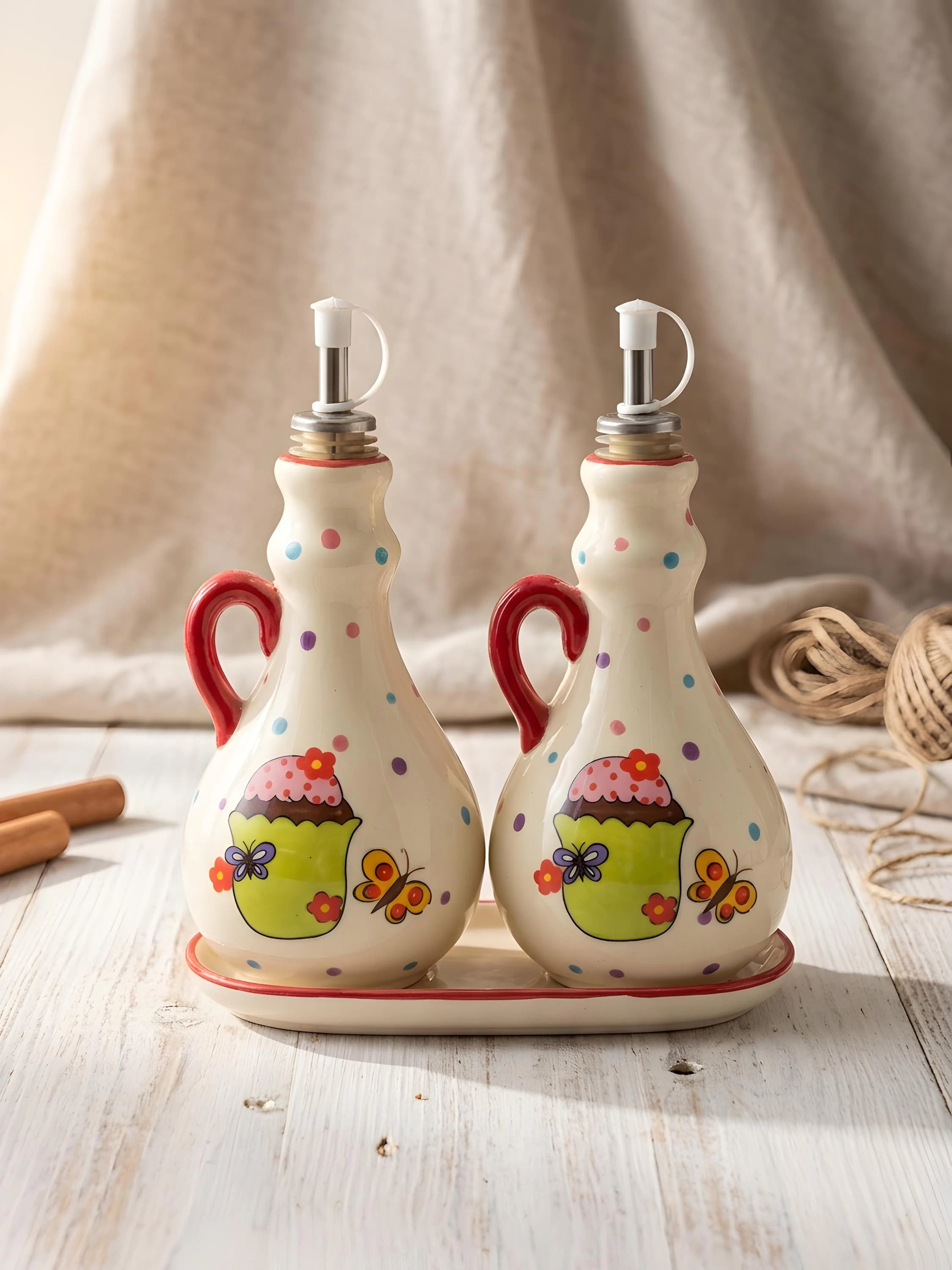 A charming set of two ceramic oil and vinegar dispensers featuring colorful polka dots, sweet cupcake illustrations, and yellow butterfly designs. Each bottle includes a vibrant red handle, a stainless steel pourer, and a white protective dust cap. The set sits on a matching oval ceramic tray. This handmade-style kitchenware adds a playful, rustic touch to any countertop or dining table.