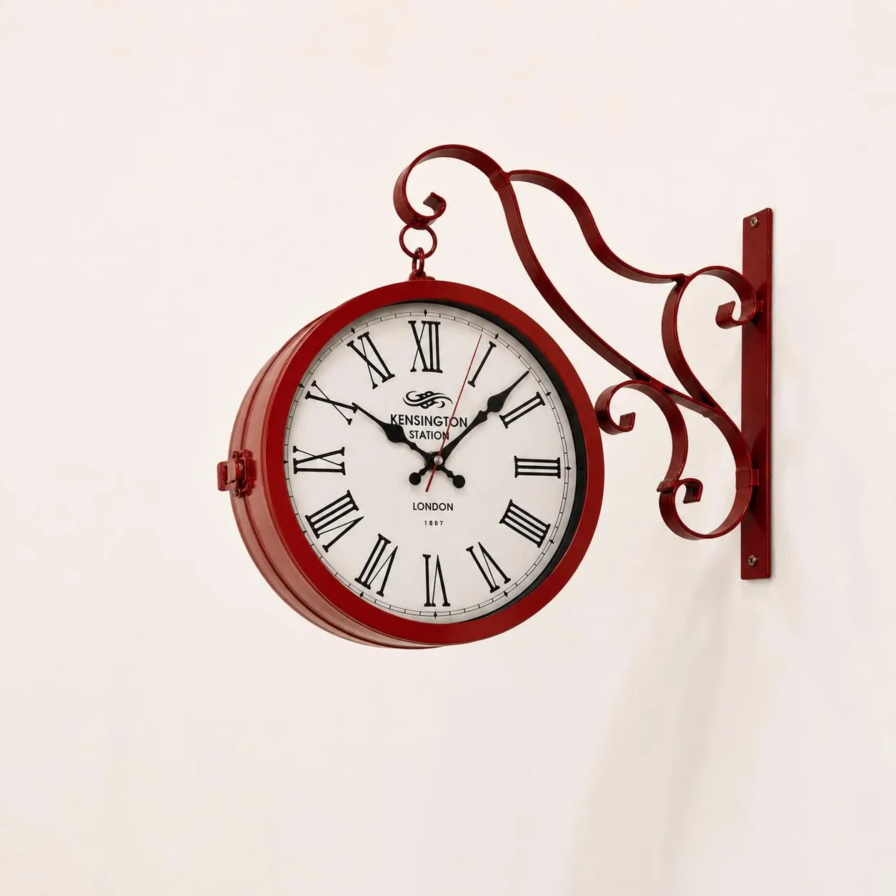 Red double-sided station wall clock with decorative scrollwork 
bracket mounted on a warm limewash living room wall. 30 cm 
Kensington Station London 1887 vintage clock displayed in cozy 
residential interior with linen armchair and dried botanicals 
nearby. Antique red railway wall clock as bold focal point in 
modern farmhouse or Victorian inspired home decor. Roman numeral 
face clearly visible.
