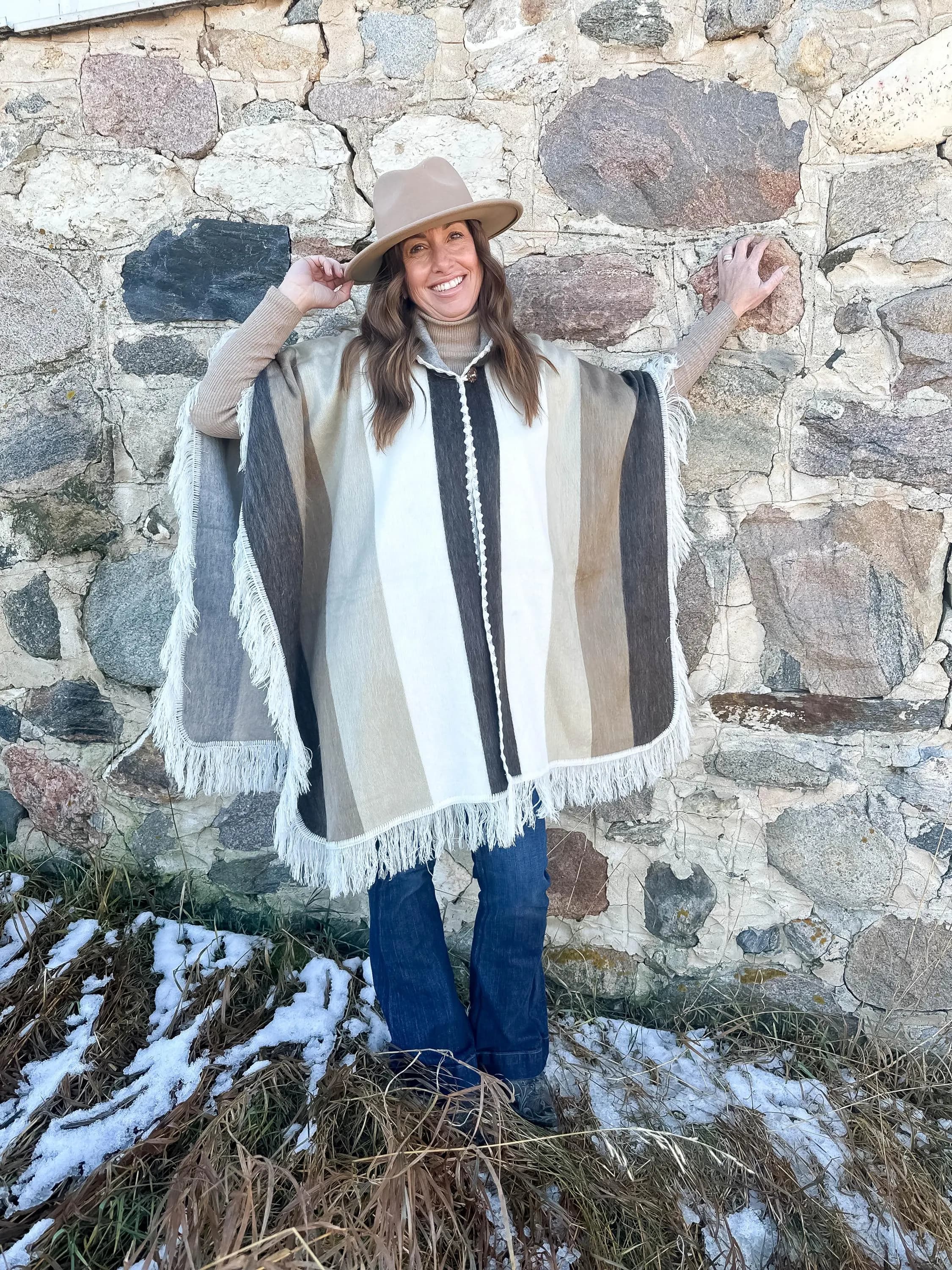 May include: A beige fedora hat and a striped poncho with fringe. The poncho features vertical stripes in shades of brown, beige, and white. The outfit is worn with blue jeans. The background is a stone wall.