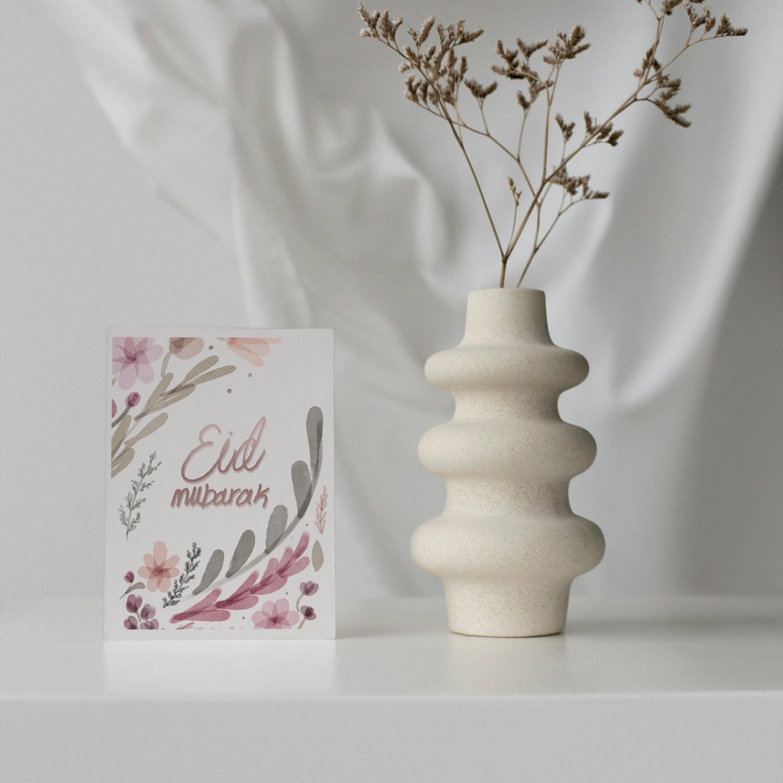 May include: A greeting card with floral watercolor design and the words "Eid Mubarak" in a script font. A beige, textured vase with a unique stacked design holds dried flowers. The card and vase are on a white surface.