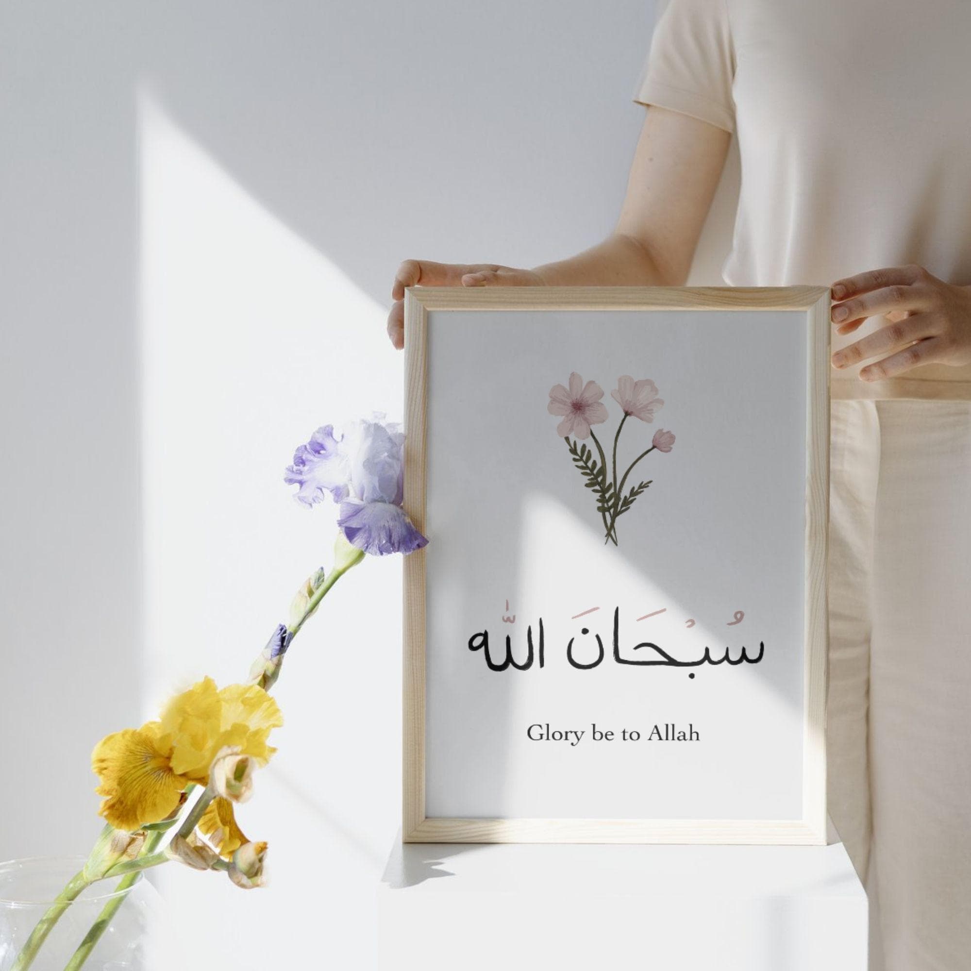 May include: A framed print with a light wood frame. The print features Arabic calligraphy and the English text "Glory be to Allah" along with a watercolor floral design. The print is held by a person.