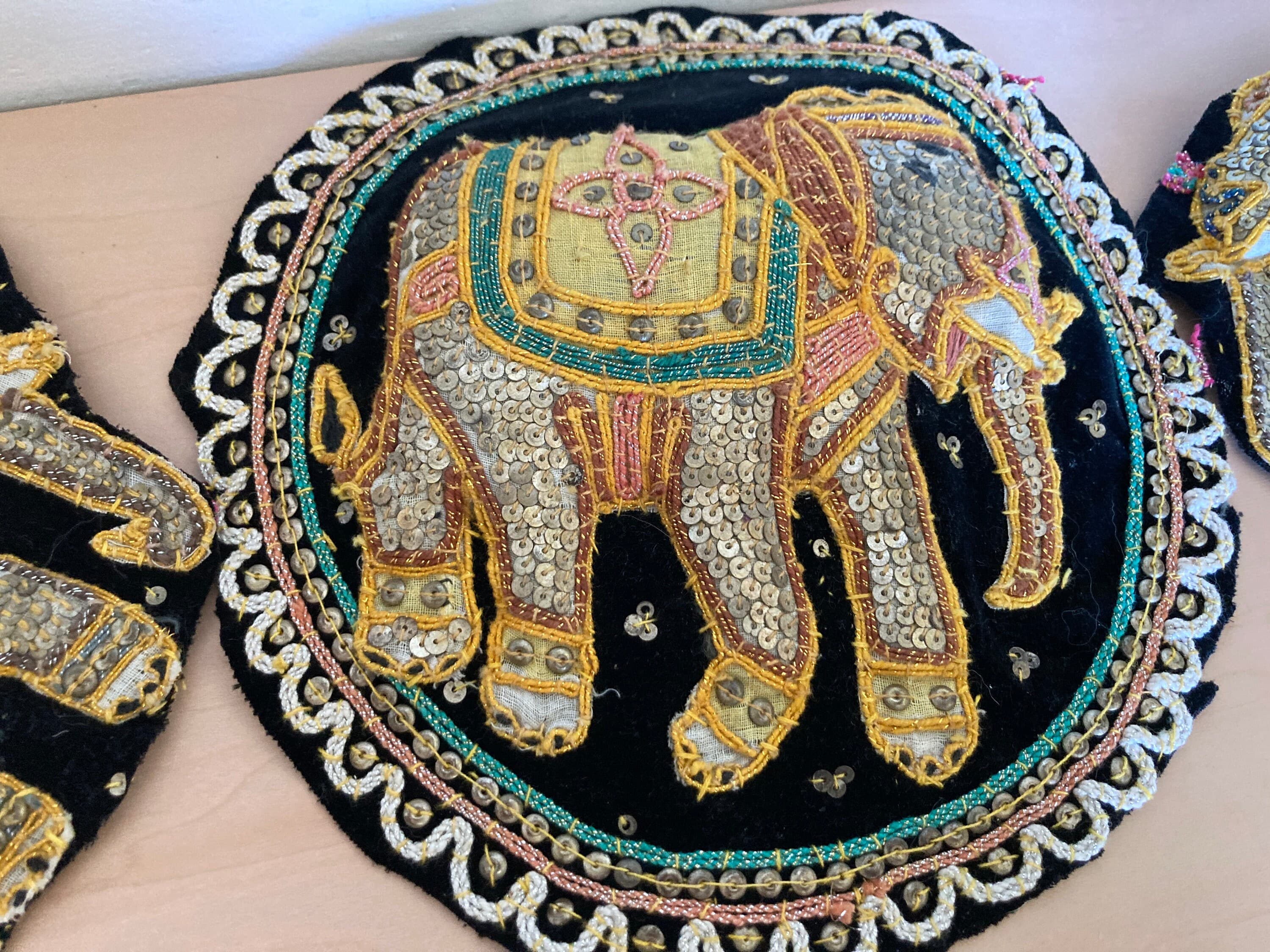 May include: Embroidered elephant applique on black fabric. The elephant is adorned with sequins, beads, and gold thread, featuring a yellow saddle and intricate detailing. The applique is surrounded by a scalloped border with gold and pink accents.