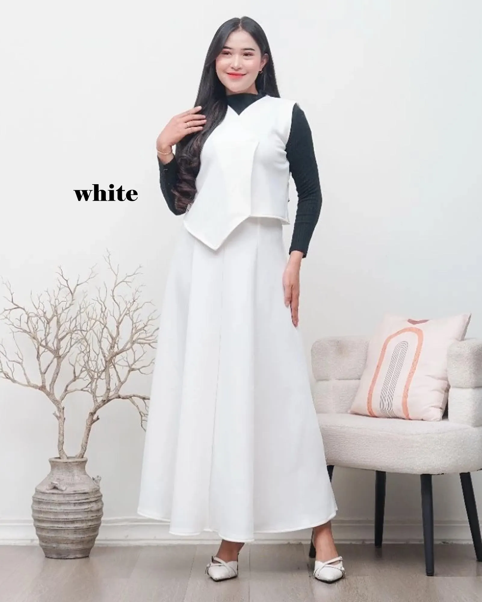 A woman wearing a sage green asymmetric scuba vest and matching A-line maxi skirt set over a white turtleneck, standing in a minimalist room