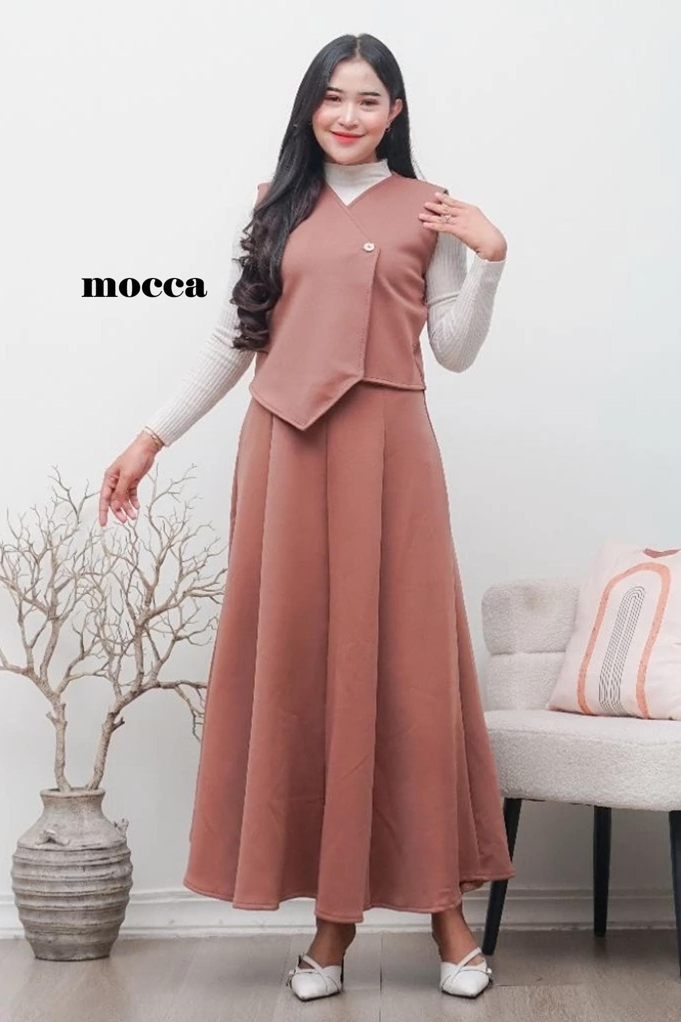 A woman wearing a sage green asymmetric scuba vest and matching A-line maxi skirt set over a white turtleneck, standing in a minimalist room