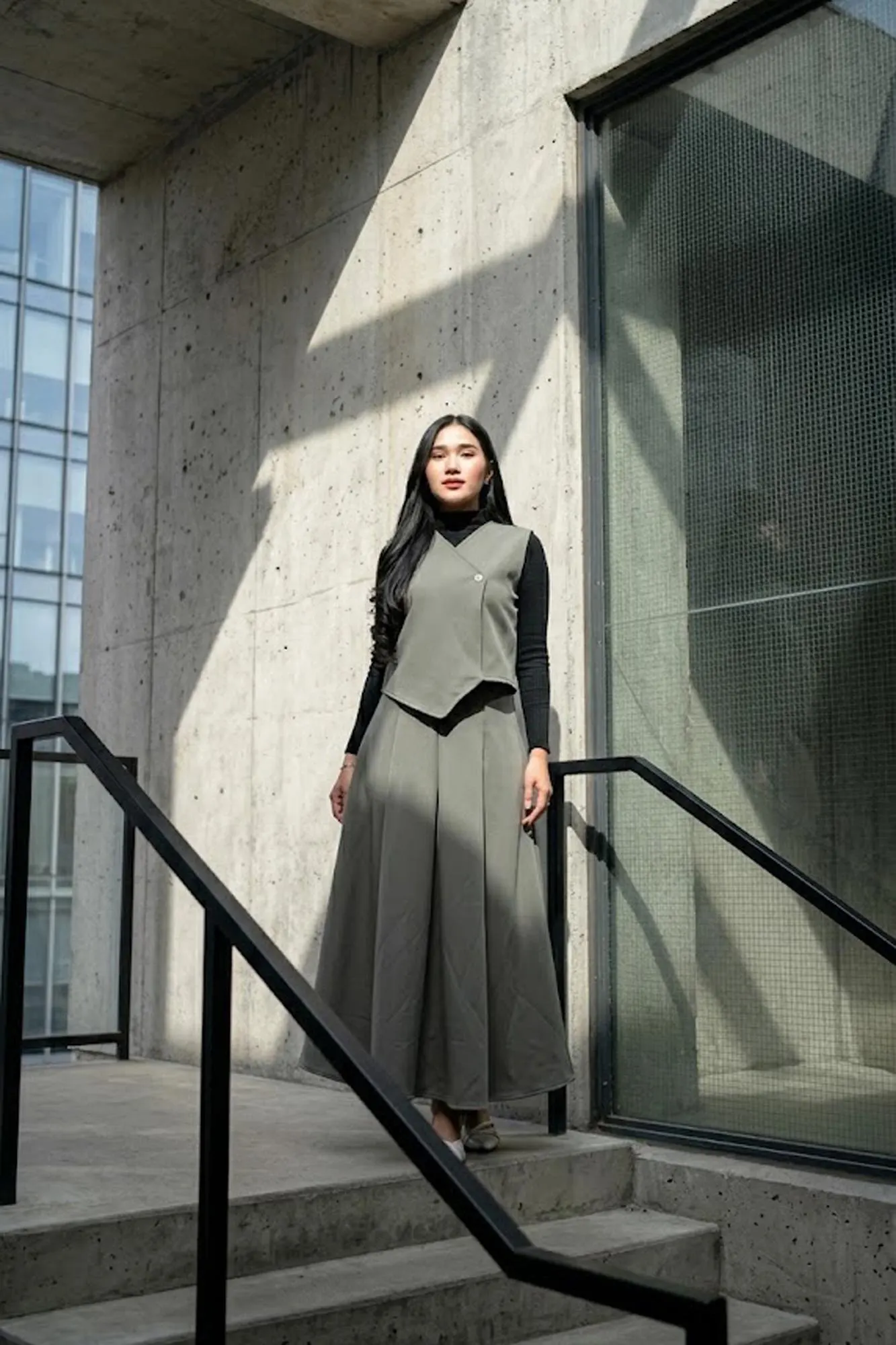 A woman wearing a sage green asymmetric scuba vest and matching A-line maxi skirt set over a white turtleneck, standing in a minimalist room