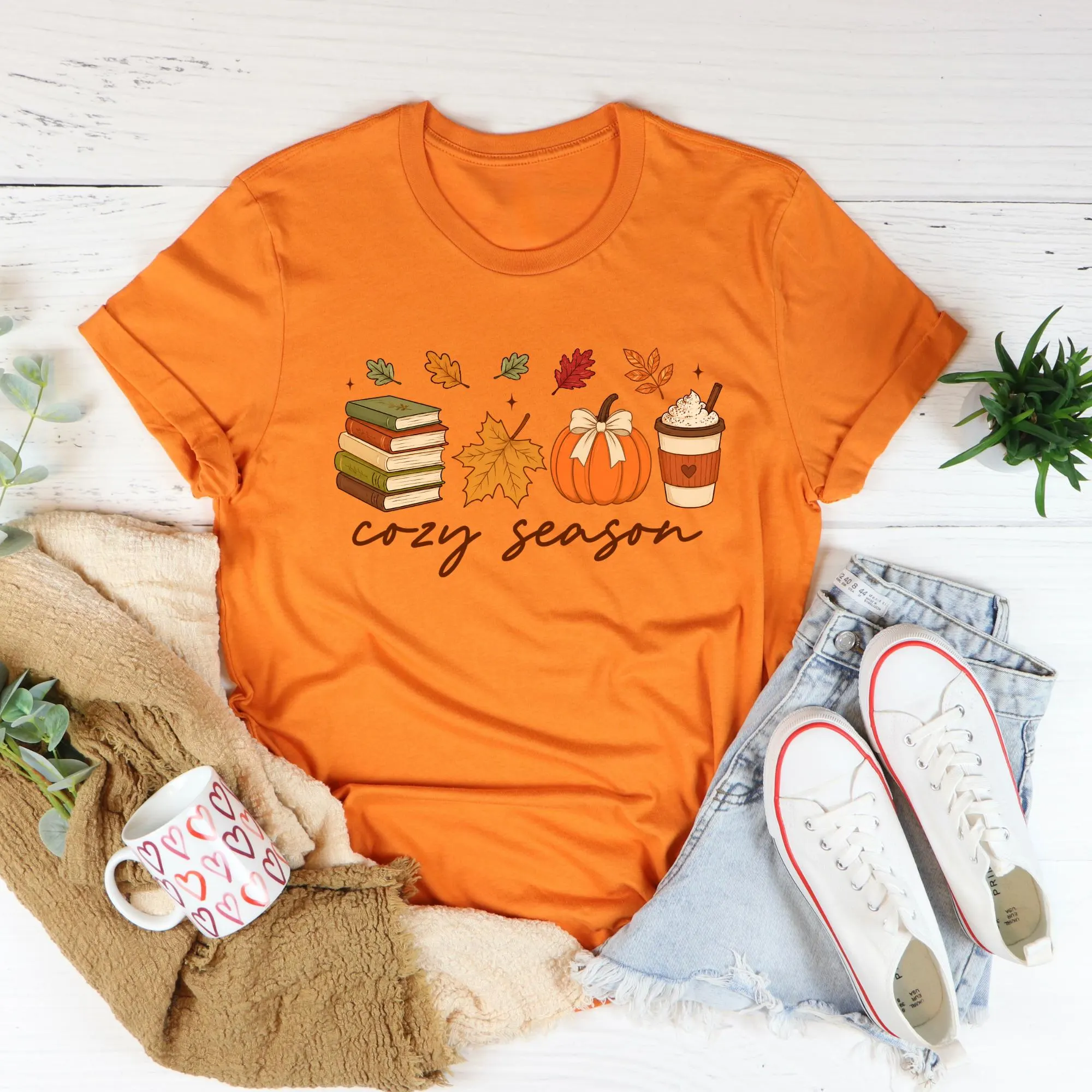 a cozy-themed orange t-shirt with illustrations of autumn elements such as books, a pumpkin, and a cup of coffee, along with the text "cozy season". The t-shirt is accompanied by a pair of denim shorts, white sneakers, and a mug with a heart design.