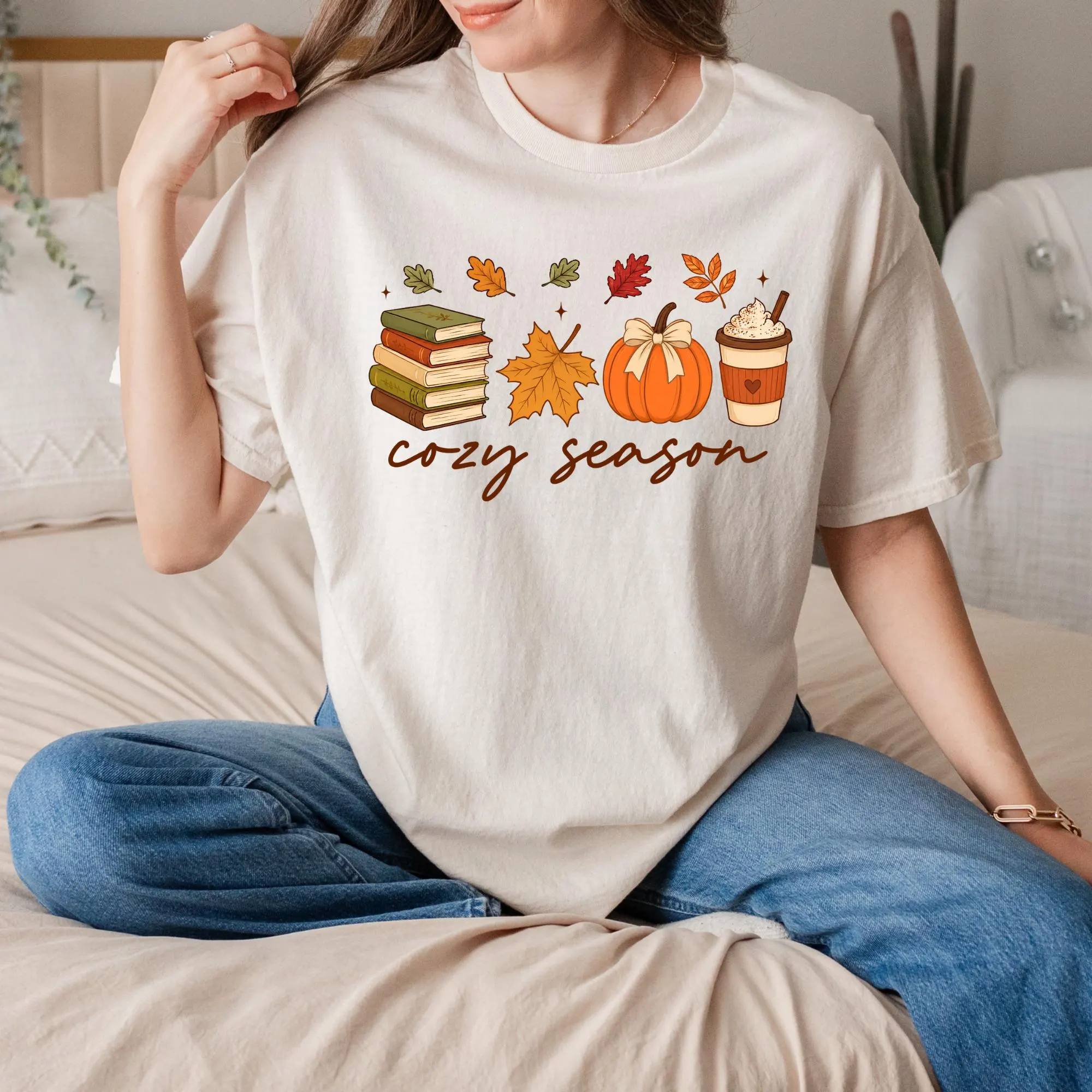 A woman wearing a white t-shirt with a graphic design of autumn-themed items, including books, a pumpkin, a cup of coffee, and a leaf, along with the text "cozy season".