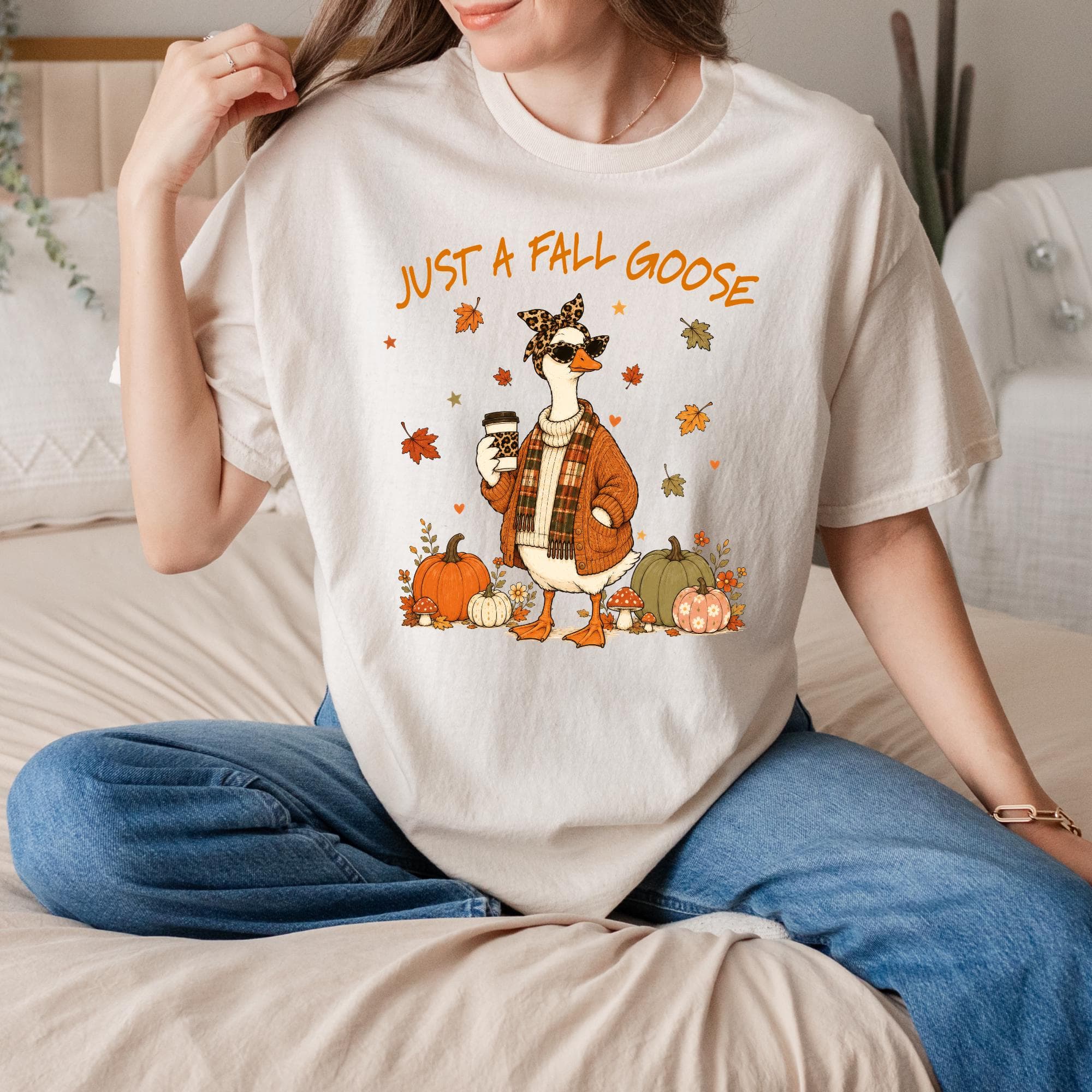 A woman wearing a white t-shirt with a graphic of a goose and autumn decorations, sitting on a bed.