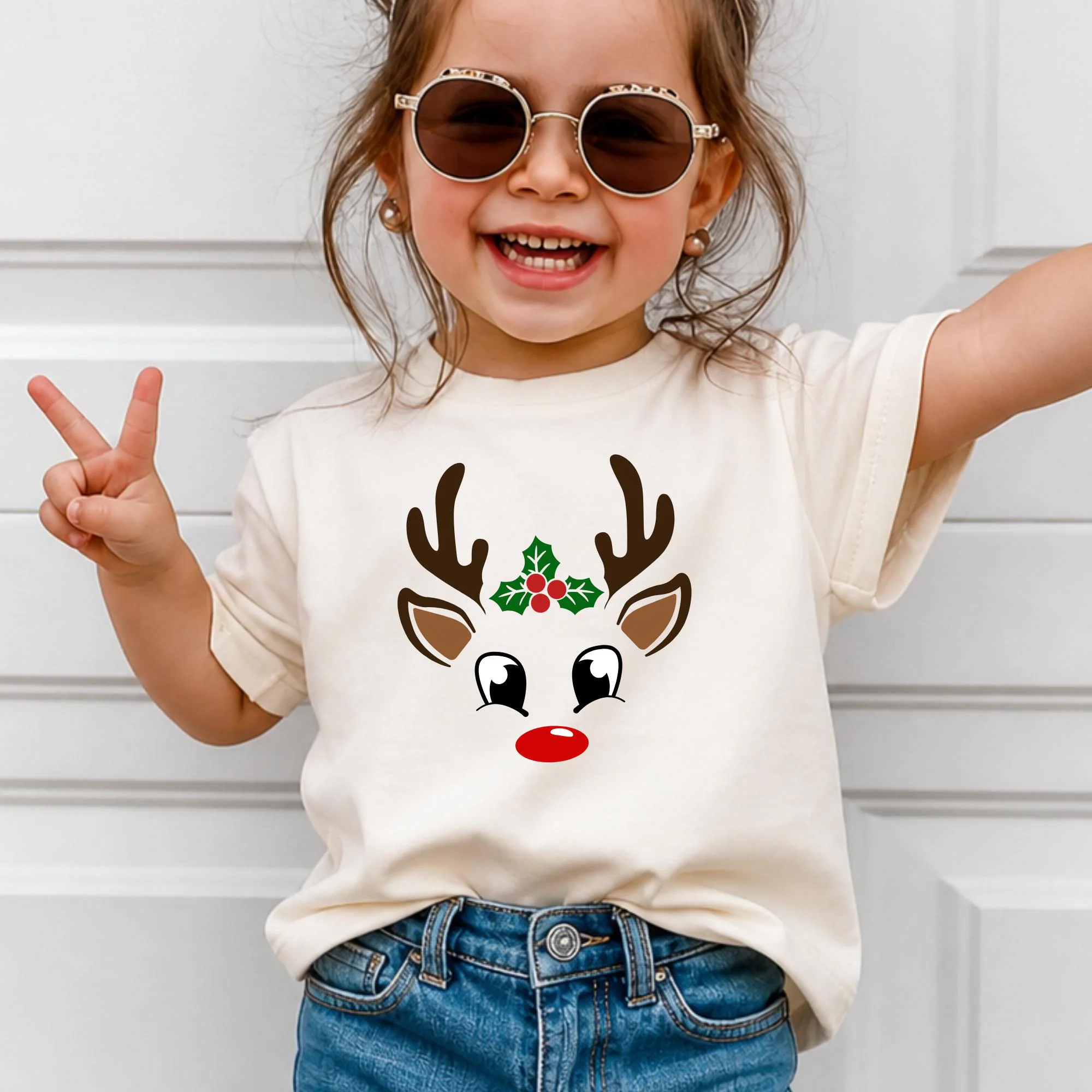 A young girl wearing a white t-shirt with a reindeer face design and sunglasses is smiling and making a peace sign.