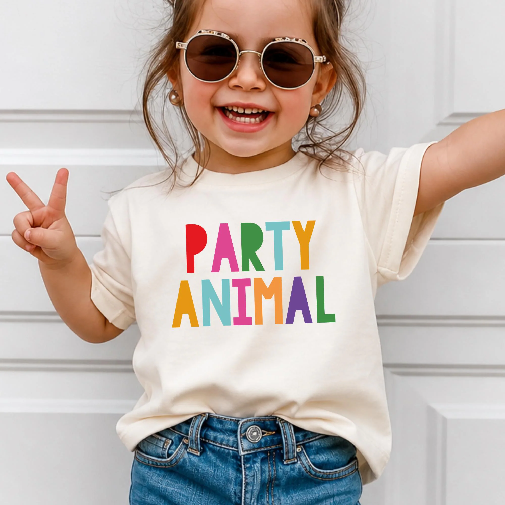 A young girl wearing sunglasses and a white t-shirt with the text "PARTY ANIMAL" is smiling and making a peace sign gesture.