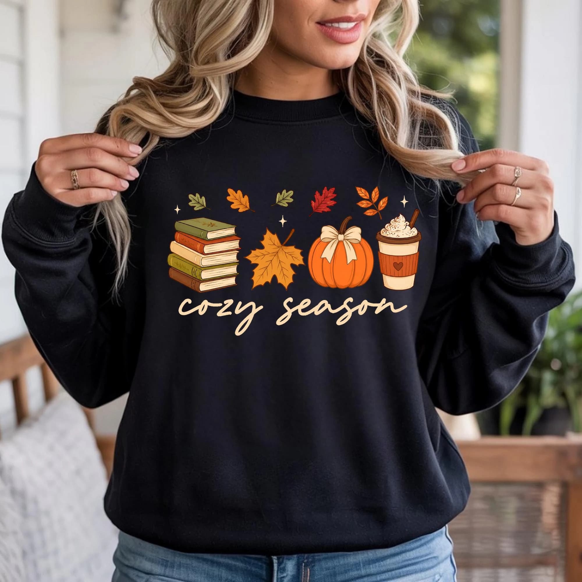 A woman wearing a black sweatshirt with a graphic design featuring autumn-themed items such as books, a pumpkin, a cup of coffee, and a scarf.