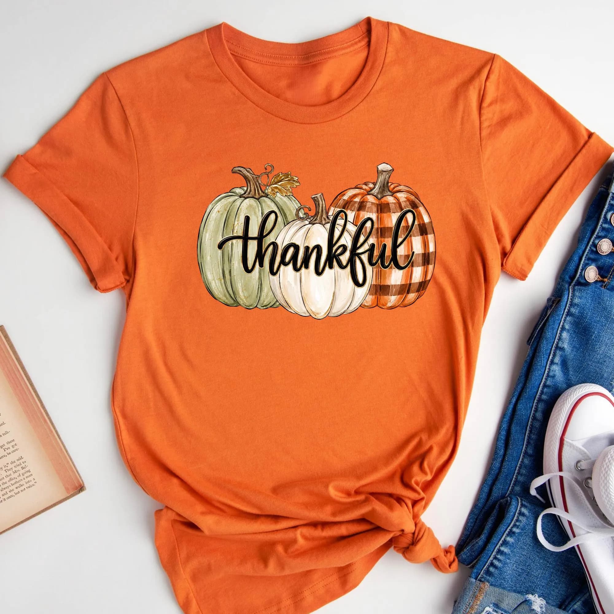 An orange t-shirt with a graphic of three pumpkins and the word "Thankful" printed on it, along with a pair of jeans and a book.