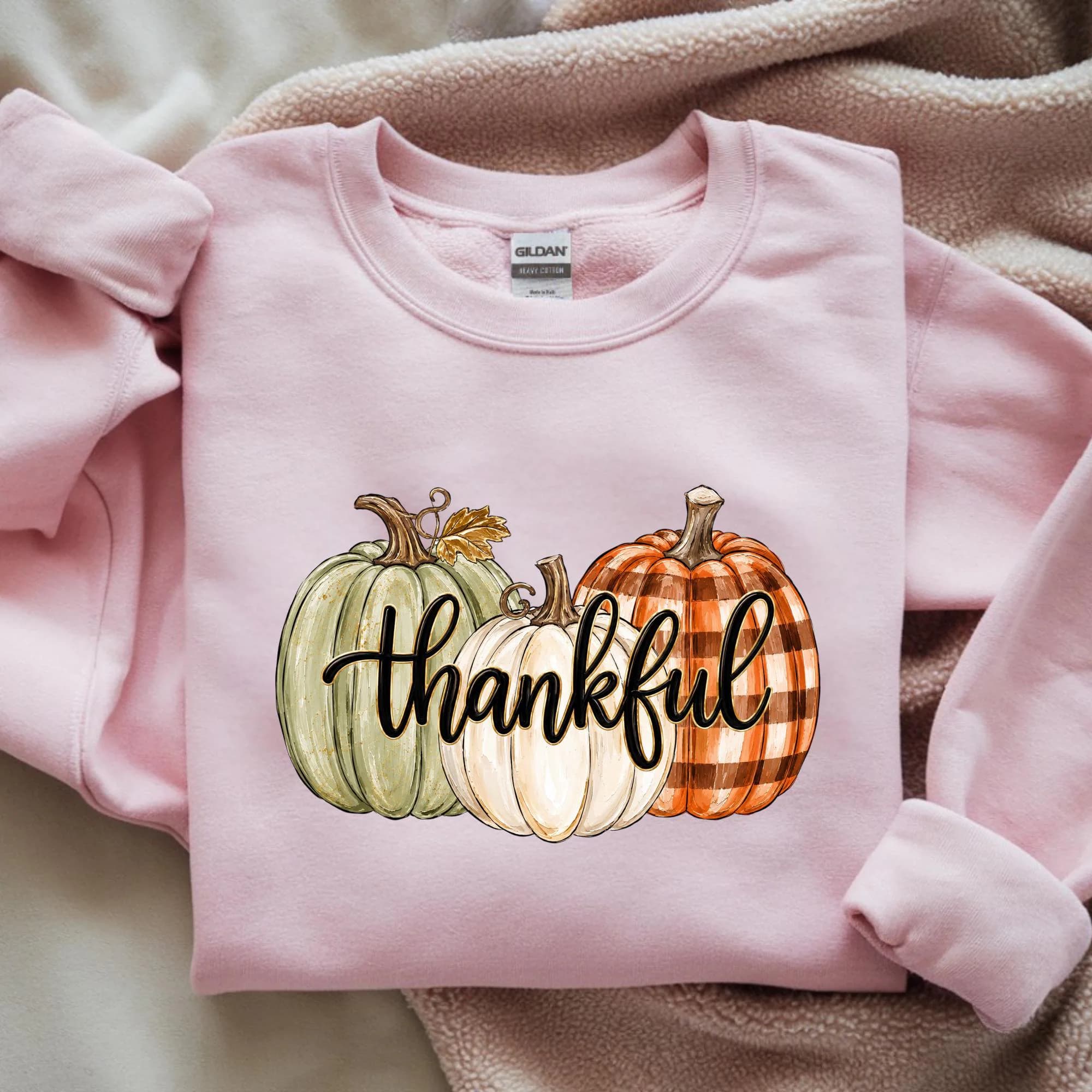 A pink sweatshirt with a graphic design of three pumpkins and the word "Thankful" printed on it.