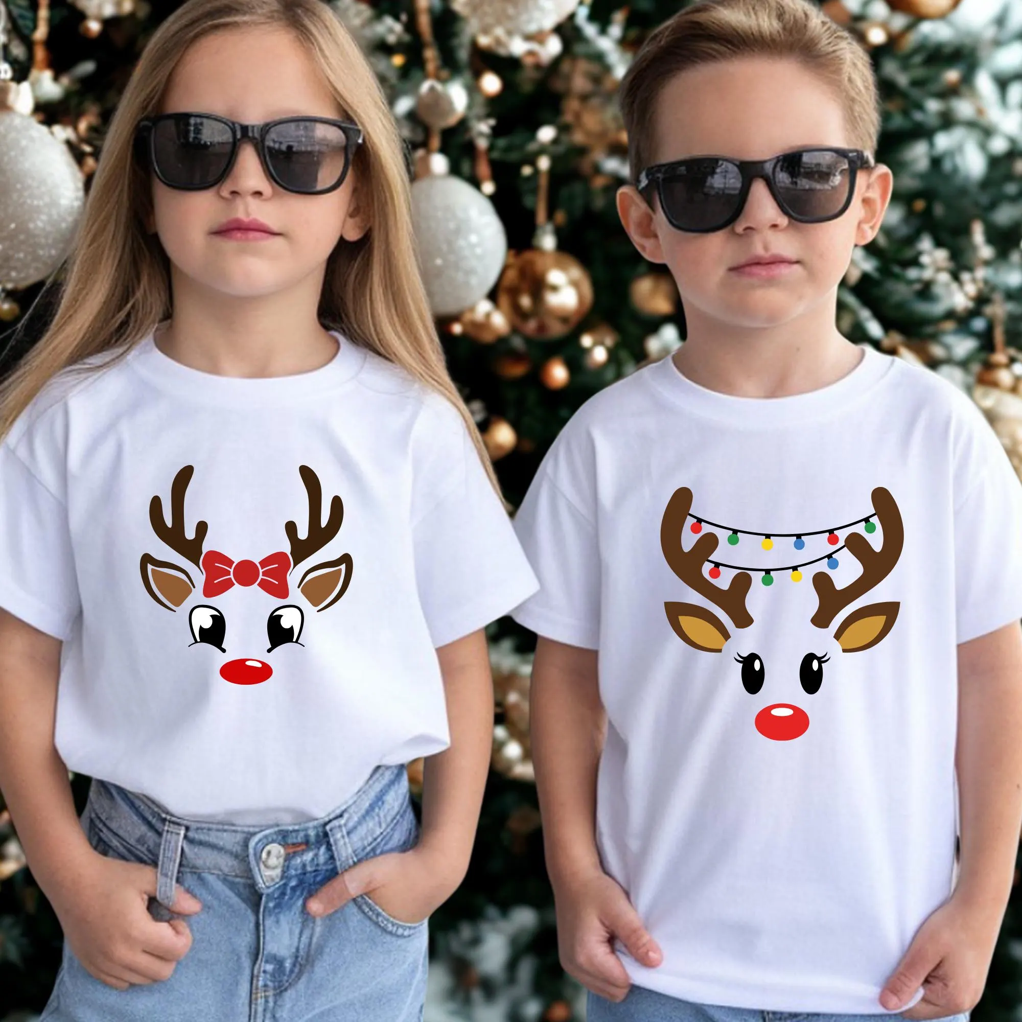 Two children wearing white t-shirts with reindeer designs stand in front of a Christmas tree.