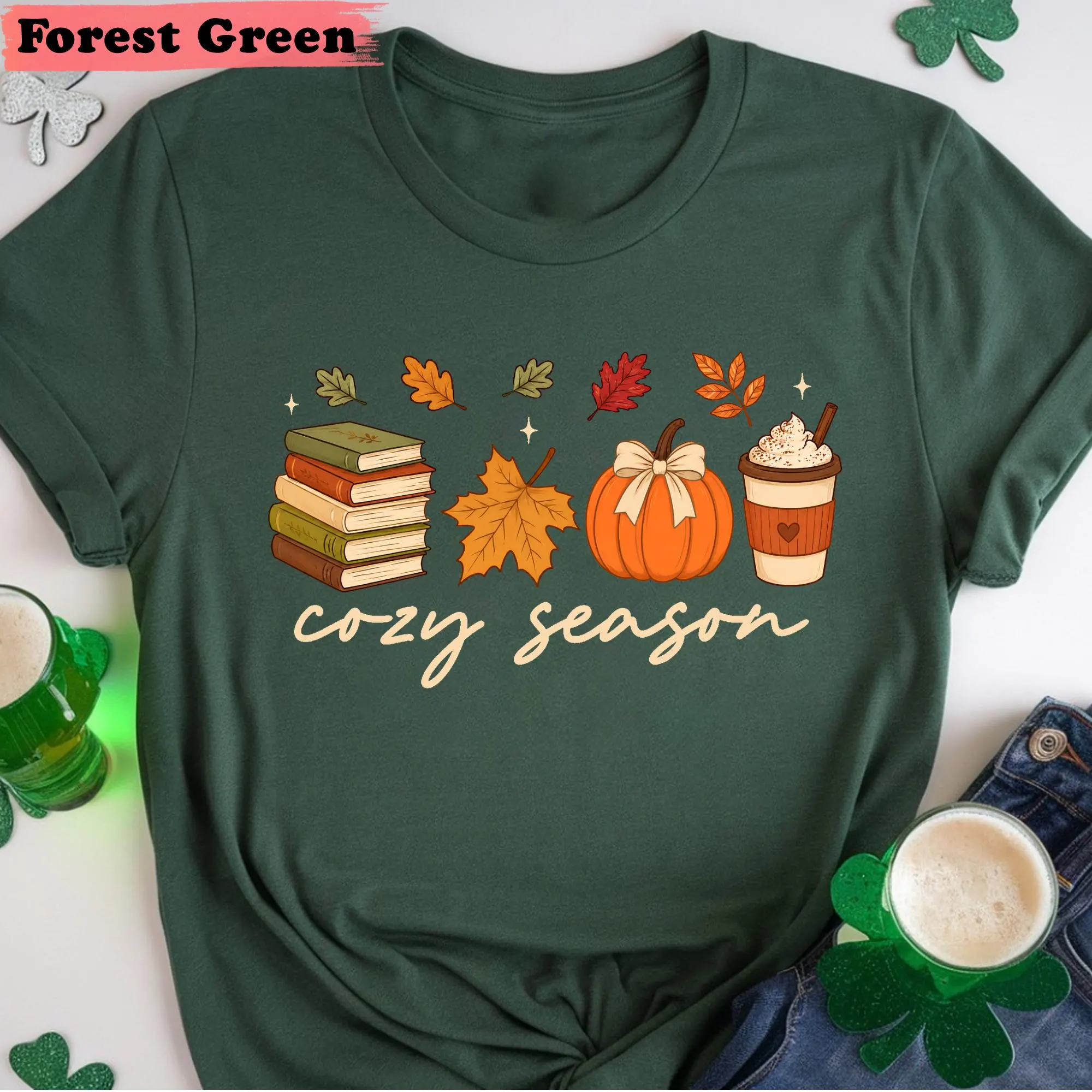 A green t-shirt with a graphic design featuring autumn-themed items such as books, a pumpkin, a cup of coffee, and a shamrock, along with the text "cozy season".