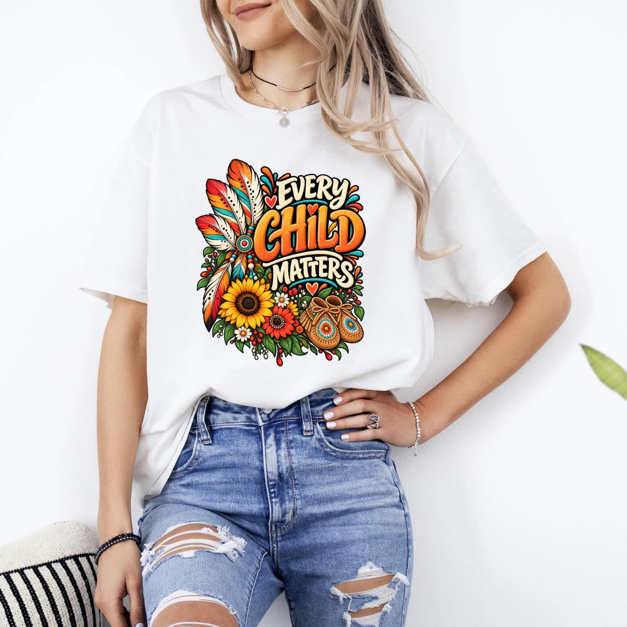 White t-shirt with colorful "Every Child Matters" text, feathers, flowers, and moccasins design.