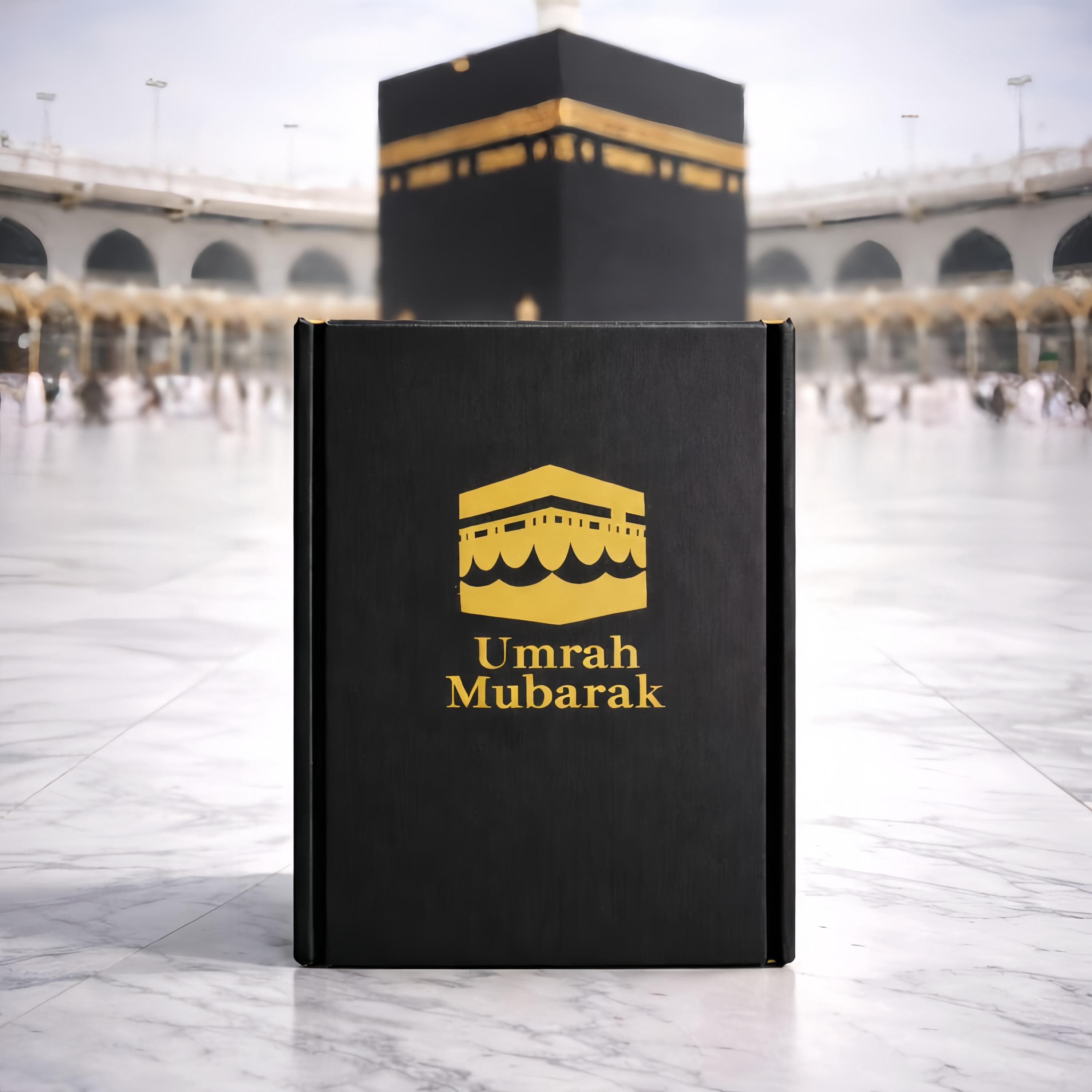 Black Umrah journal displayed upright on a marble floor, featuring a gold Kaaba icon and the words “Umrah Mubarak” on the cover. The journal is shown with the Kaaba in the background at Masjid al-Haram, highlighting the b