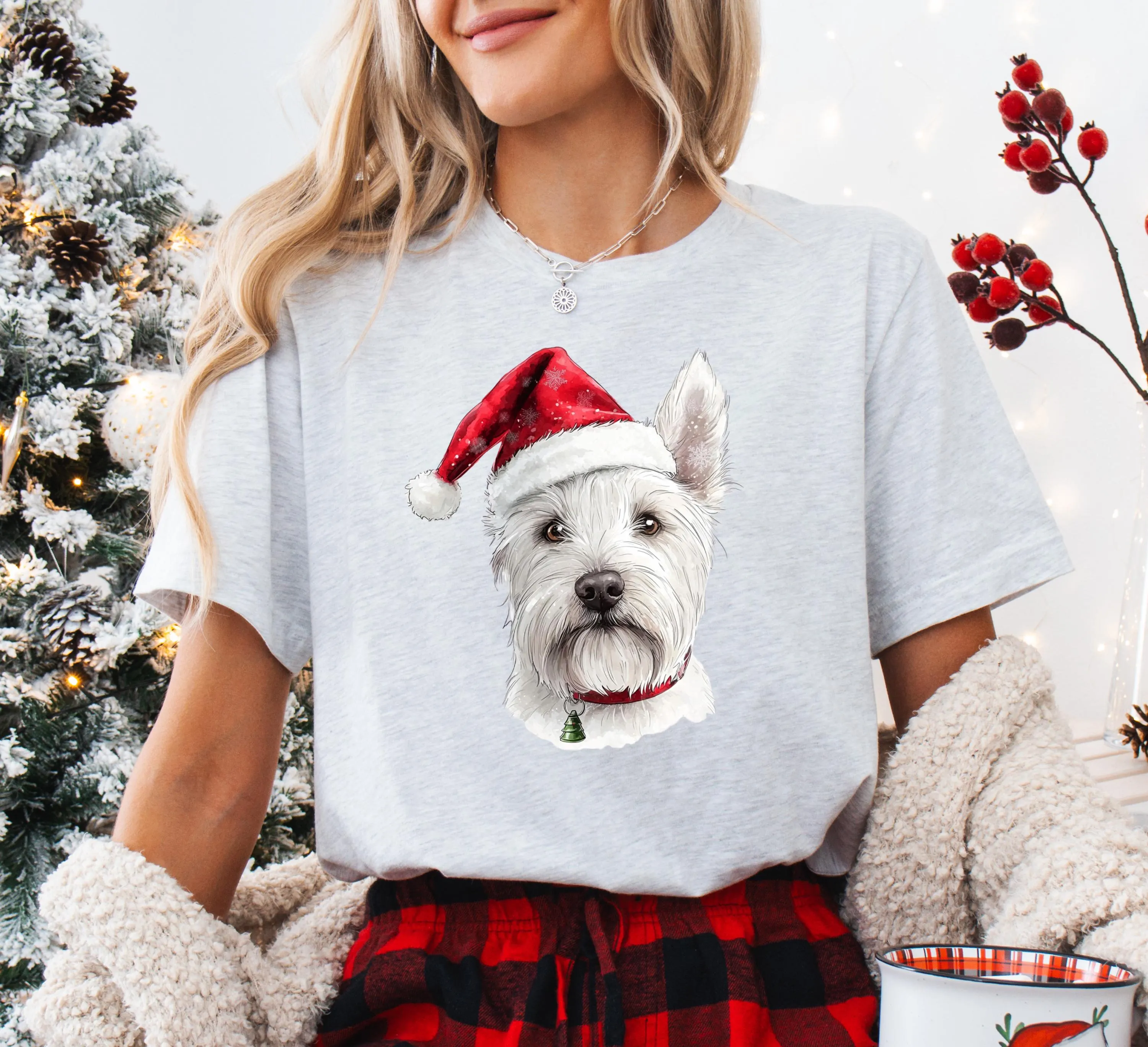 A woman wearing a gray t-shirt with a white Westie dog wearing a Santa hat, standing in front of a Christmas tree.