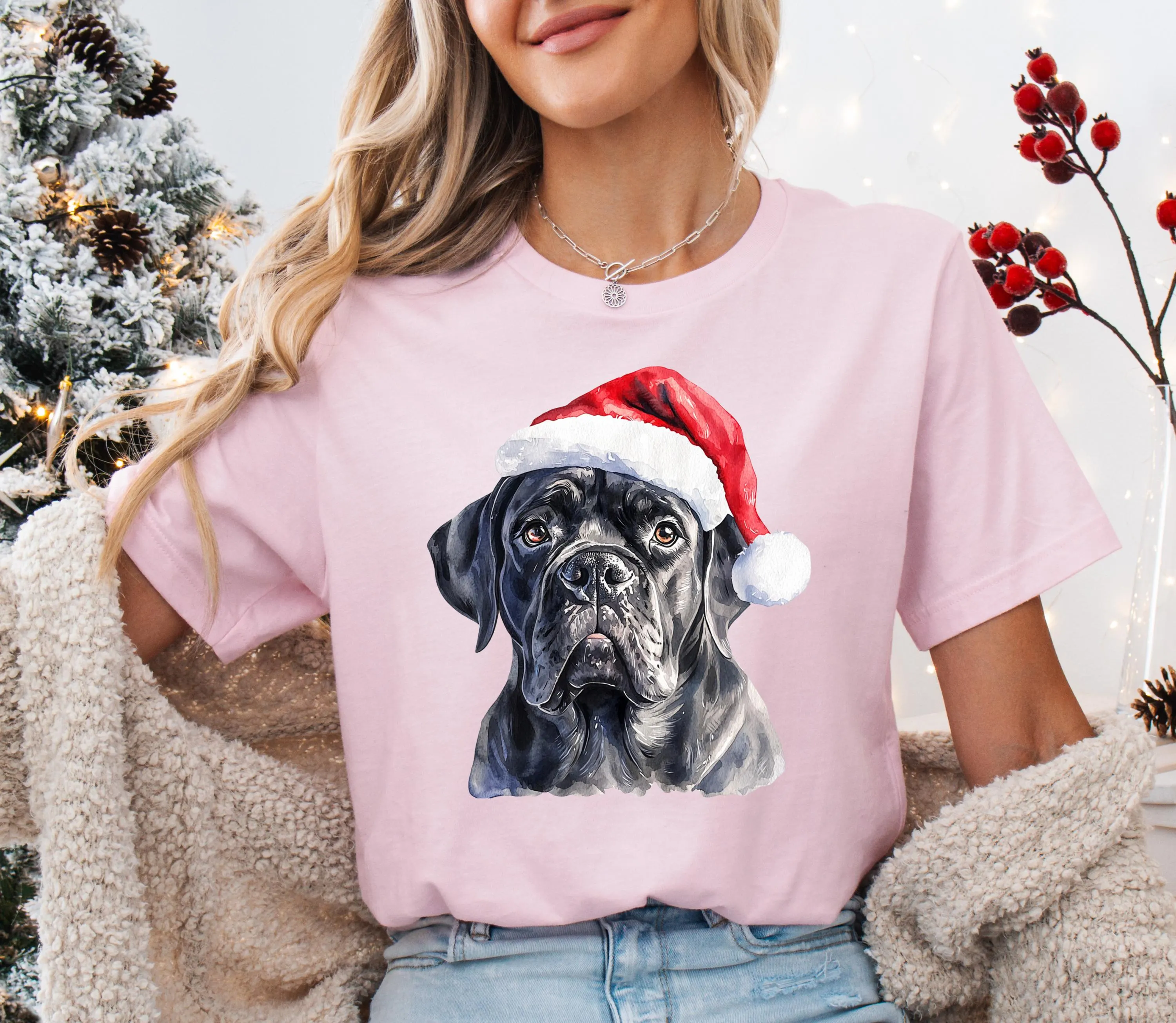A woman wearing a pink t-shirt with a black dog wearing a red Santa hat.