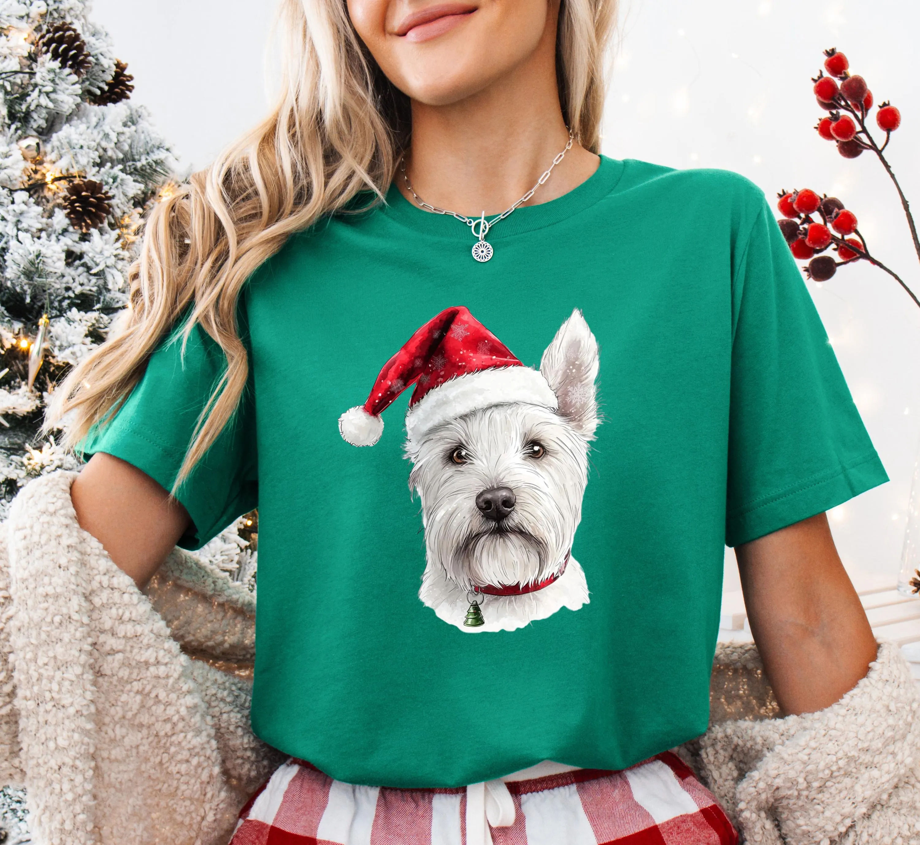 A woman wearing a green t-shirt with a white dog wearing a Santa hat printed on it, standing in front of a Christmas tree.