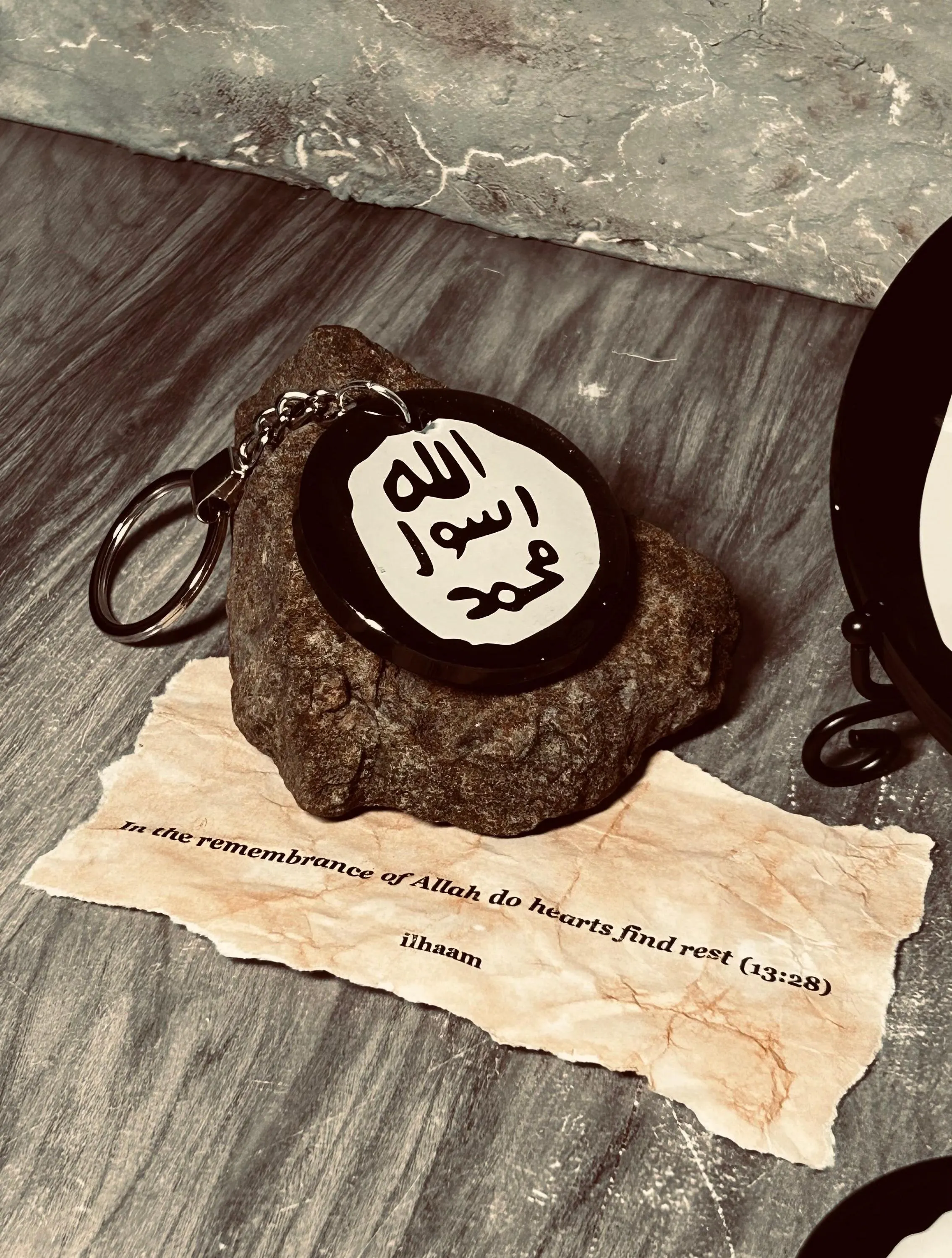 A small round black keyring with white Arabic calligraphy rests on a dark stone base, placed on a textured surface with a beige paper note underneath and part of a matching circular item visible at the right edge.