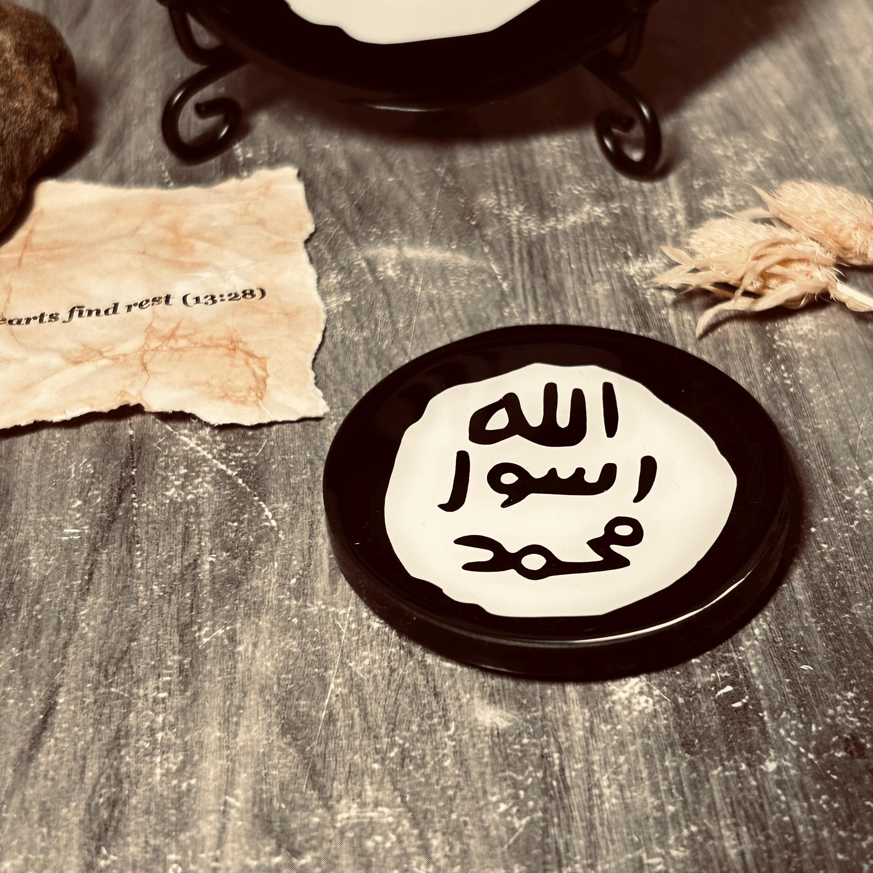 A round black and white Islamic Fridge magnet with Arabic calligraphy reading 'Allah Rasool Muhammad' resting on a rustic grey wooden surface with styled props.