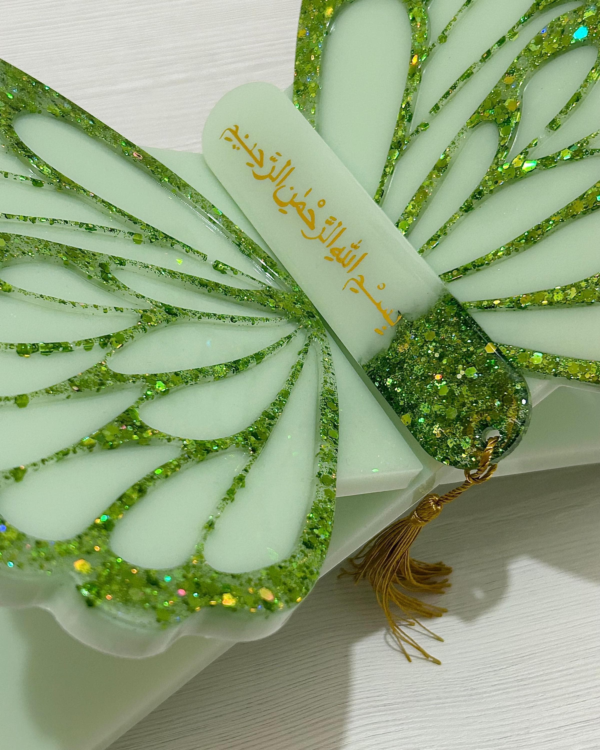 Part of a butterfly design Quran stand in sage green and green glitter detailed wings with a matching oval Quran bookmark with Bismillah in gold text and a gold tassel on a wood grain textured background