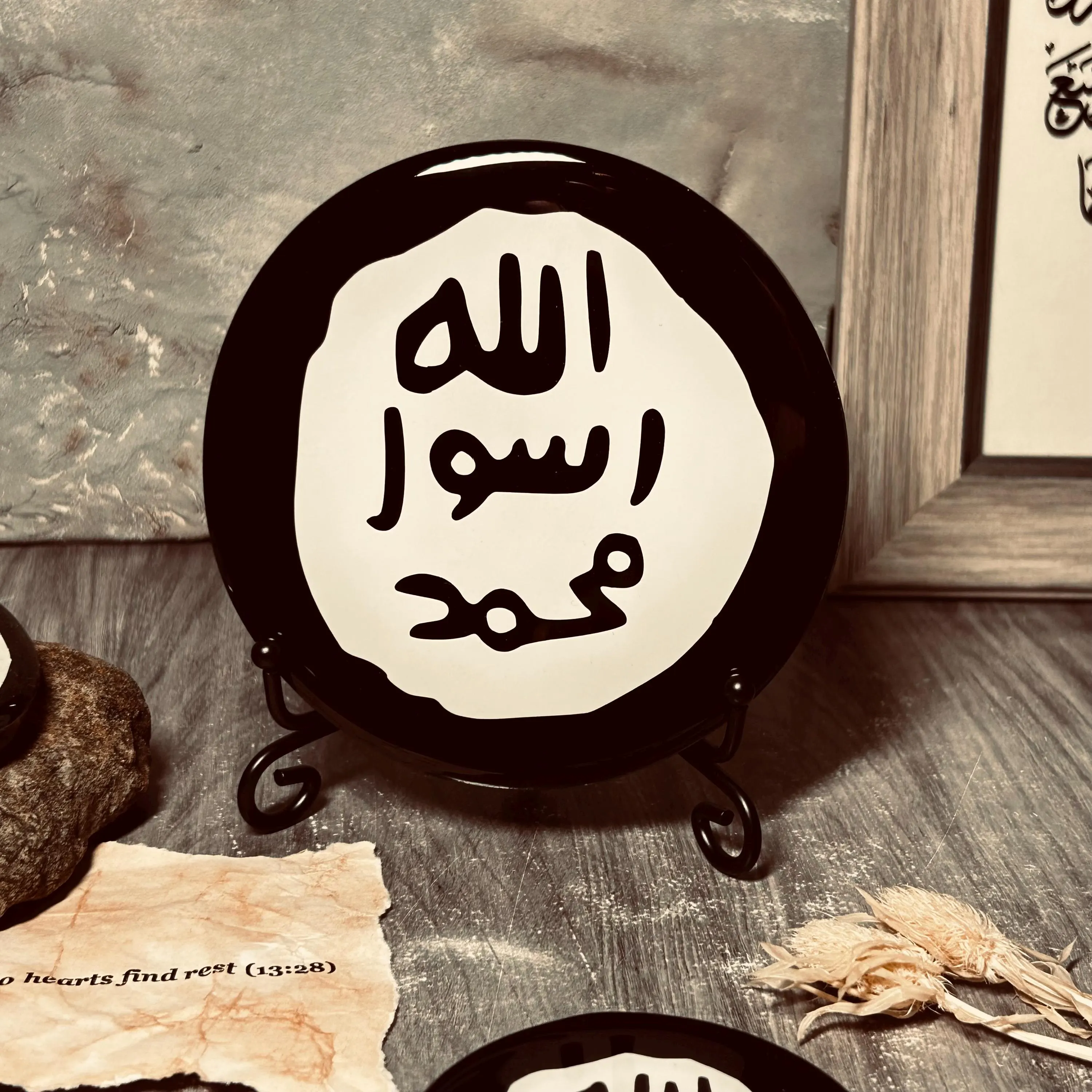 A round black and white Islamic plaque featuring Arabic calligraphy that reads 'Allah Rasool Muhammad, displayed on a small black stand on a rustic wooden surface.