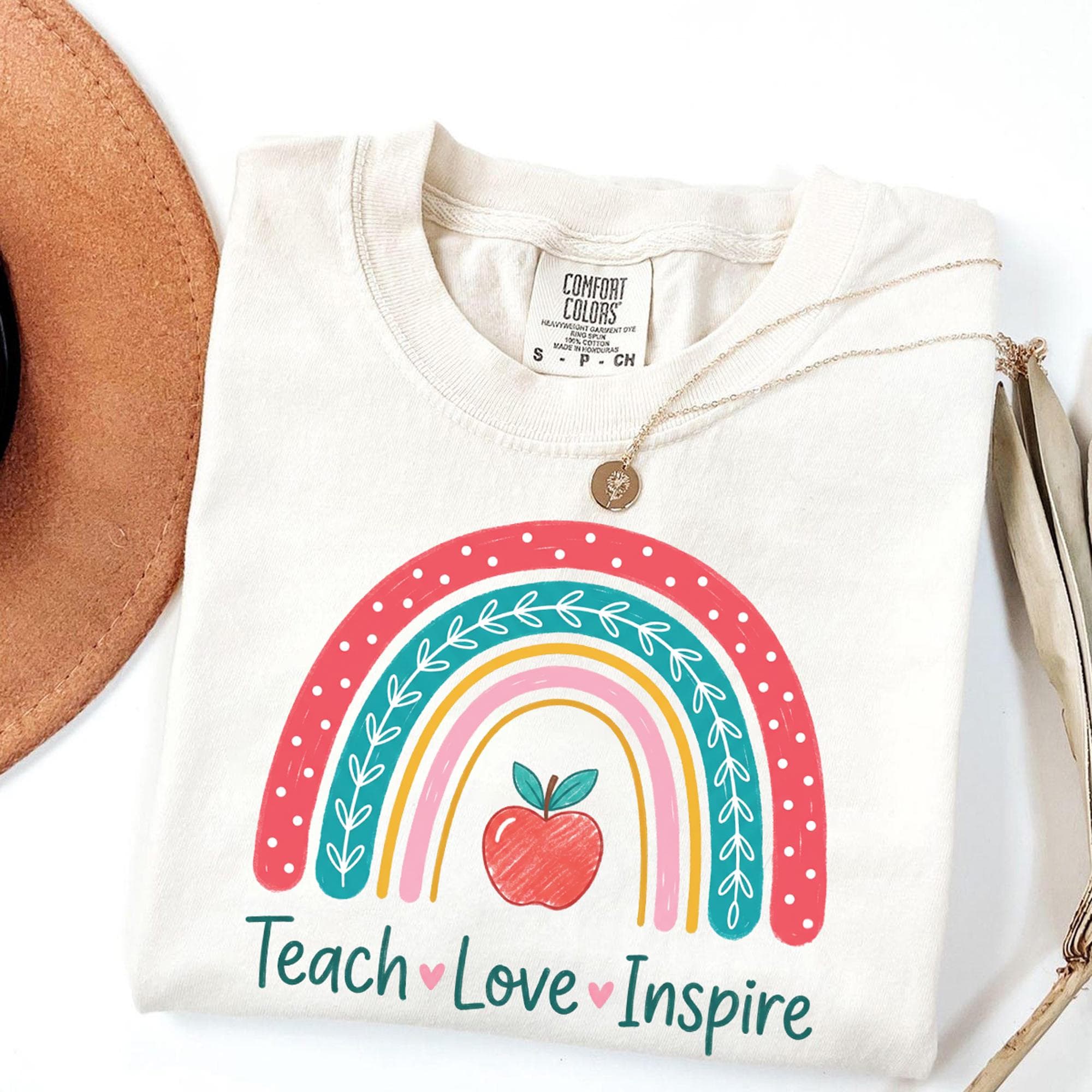 A white t-shirt with a colorful graphic design, including a rainbow, apple, and the text "Teach Love Inspire".