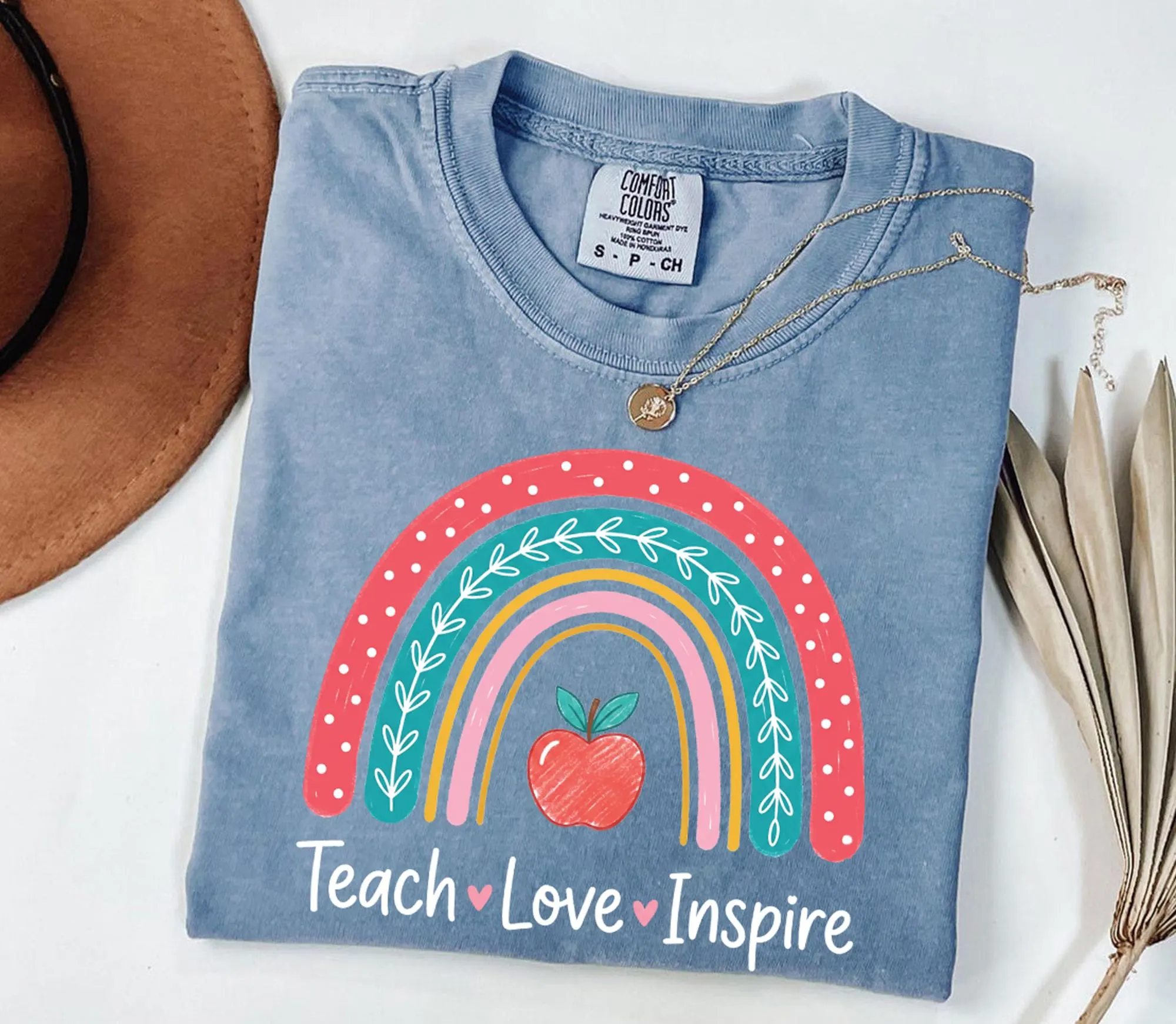 A blue t-shirt with a colorful graphic design, including a rainbow and an apple, along with the text "Teach Love Inspire" printed on it.