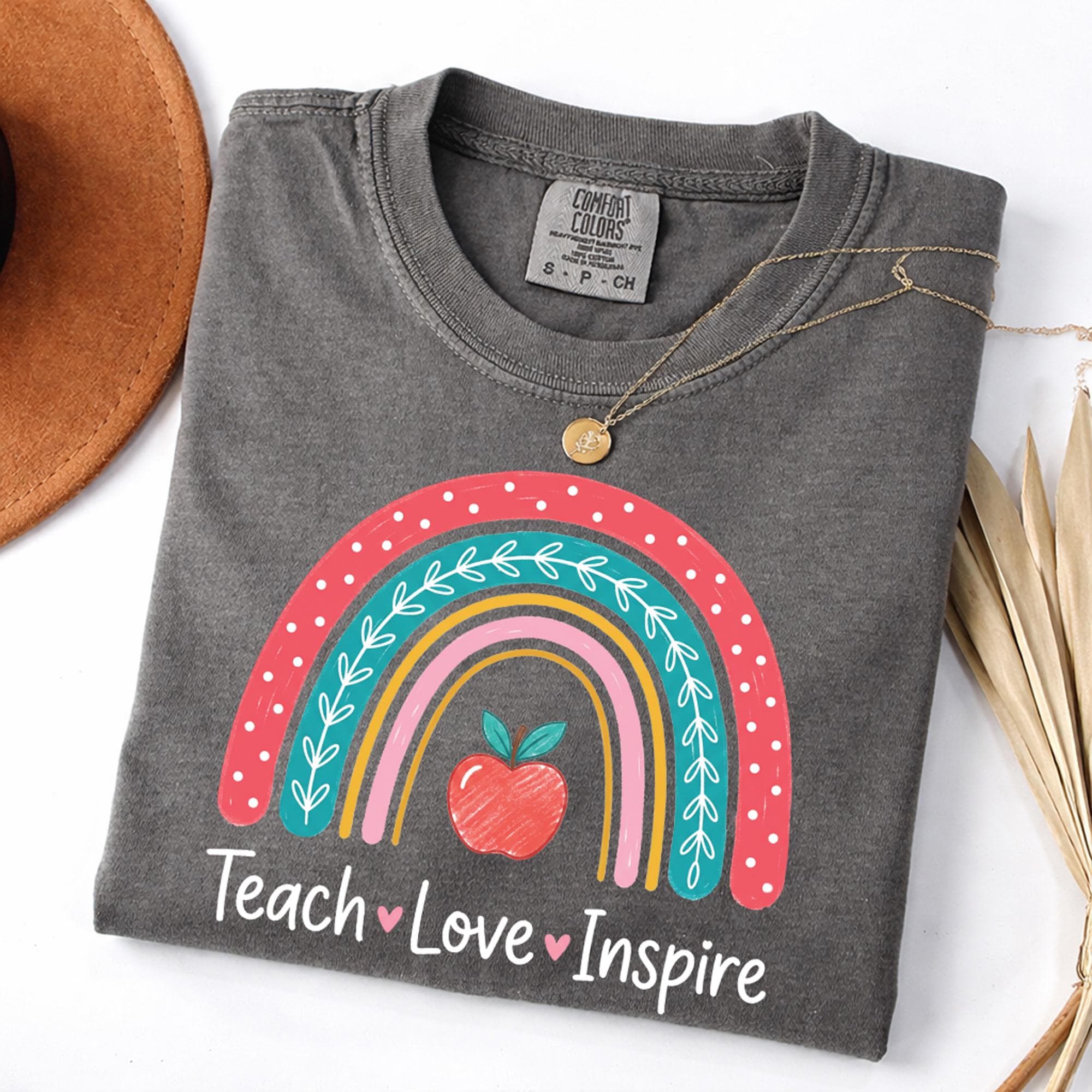 a gray t-shirt with a colorful graphic design, including a rainbow and an apple, along with the text "Teach Love Inspire".