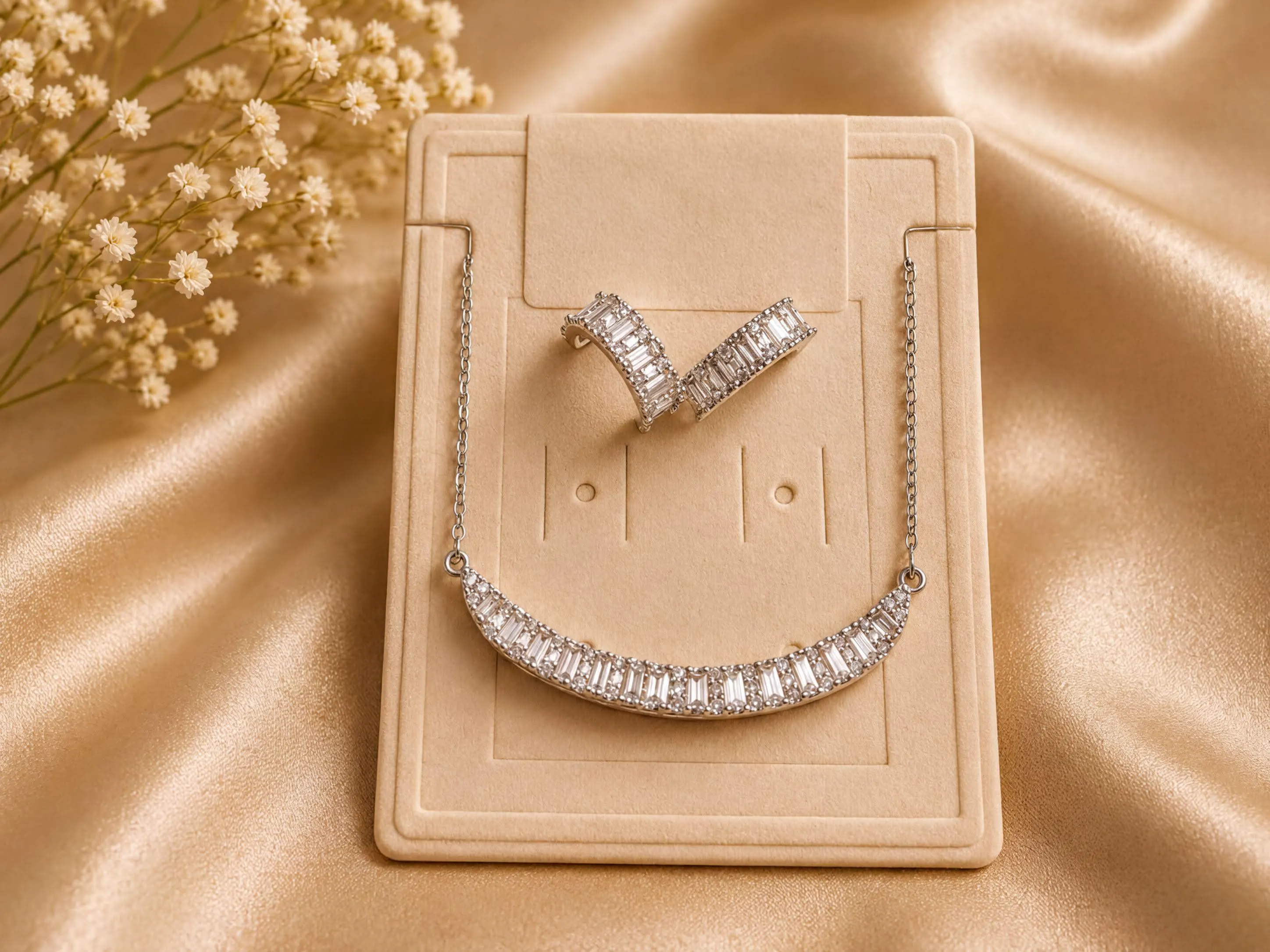 A necklace and earrings are displayed on a beige background, with a small floral arrangement in the background.