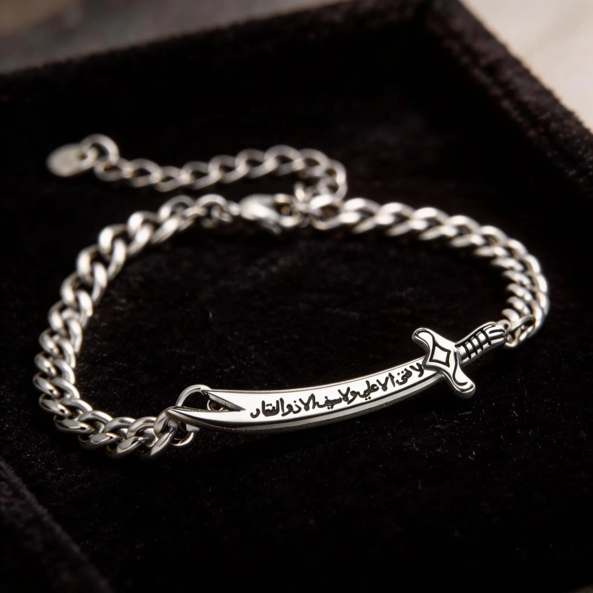 A silver-colored curb chain bracelet featuring a sword pendant inscribed with Arabic text, resting in a black velvet-lined box.