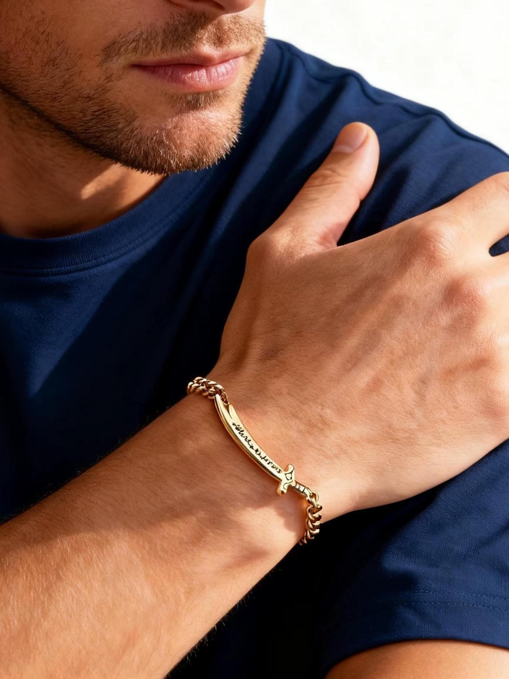 Close-up of a person wearing a gold sword-shaped curb chain bracelet on their wrist and a blue t-shirt.