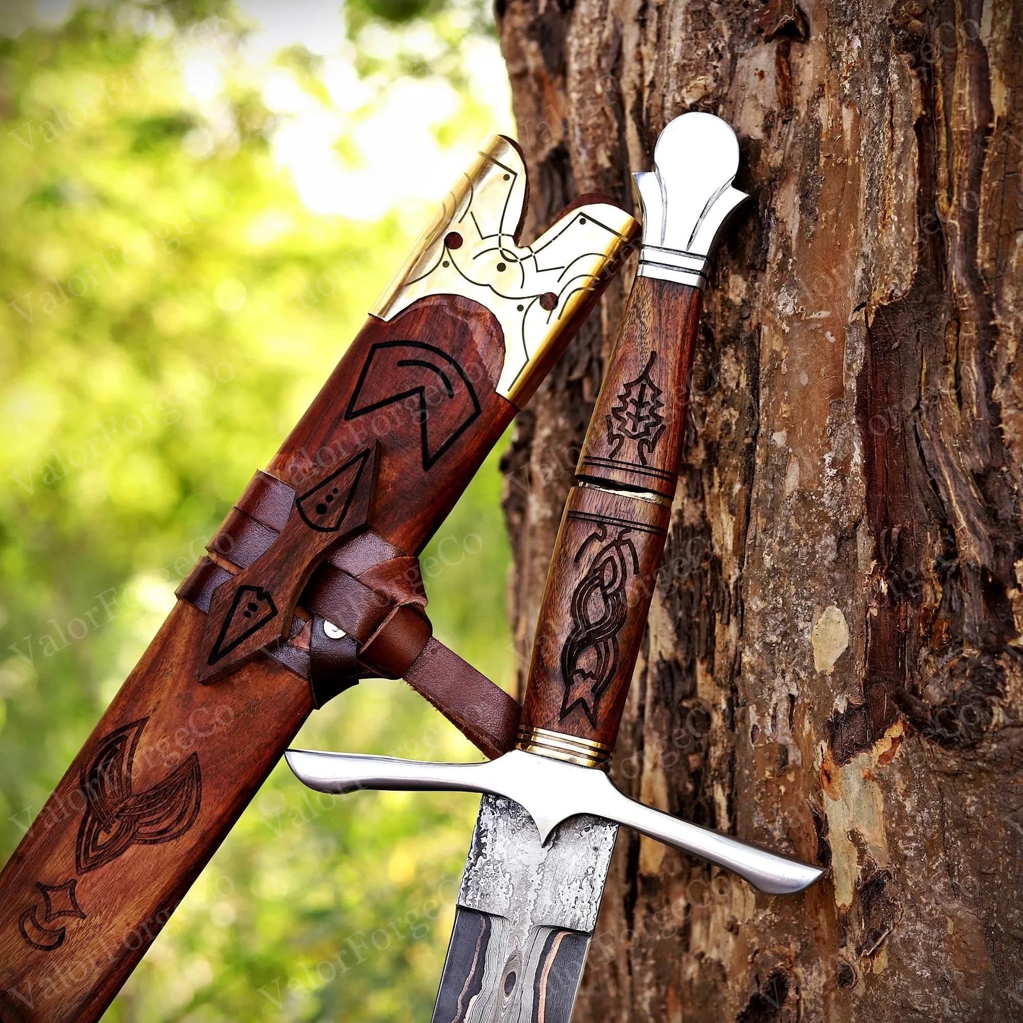 A sword with a patterned steel blade, a wooden hilt featuring intricate carvings, and a silver-coloured pommel. The matching wooden scabbard displays decorative brass accents and dark etched designs, secured by brown leather straps.