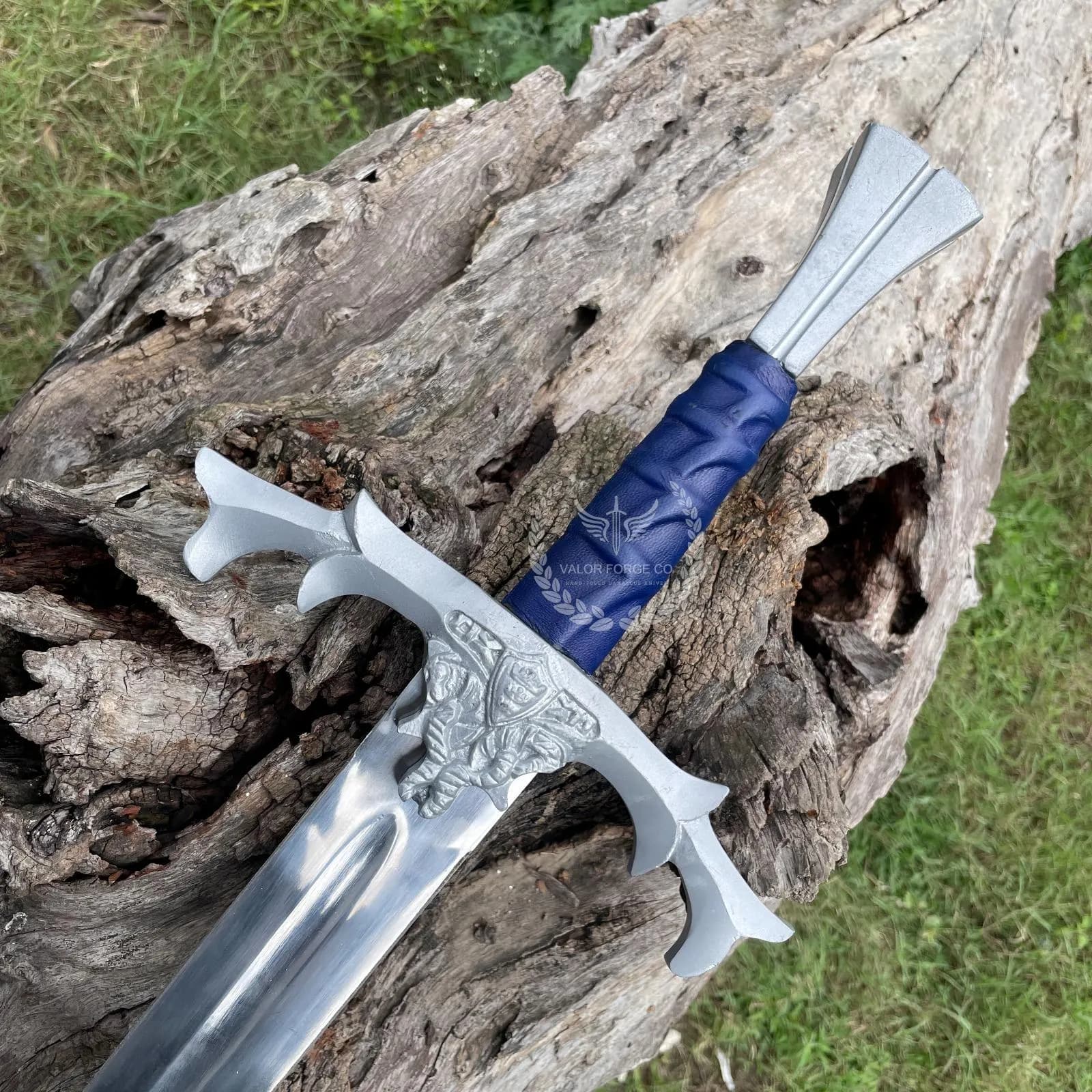 Close-up of the decorative sword resting on a tree trunk, highlighting its ornate guard, blue handle, and silver-colored blade.