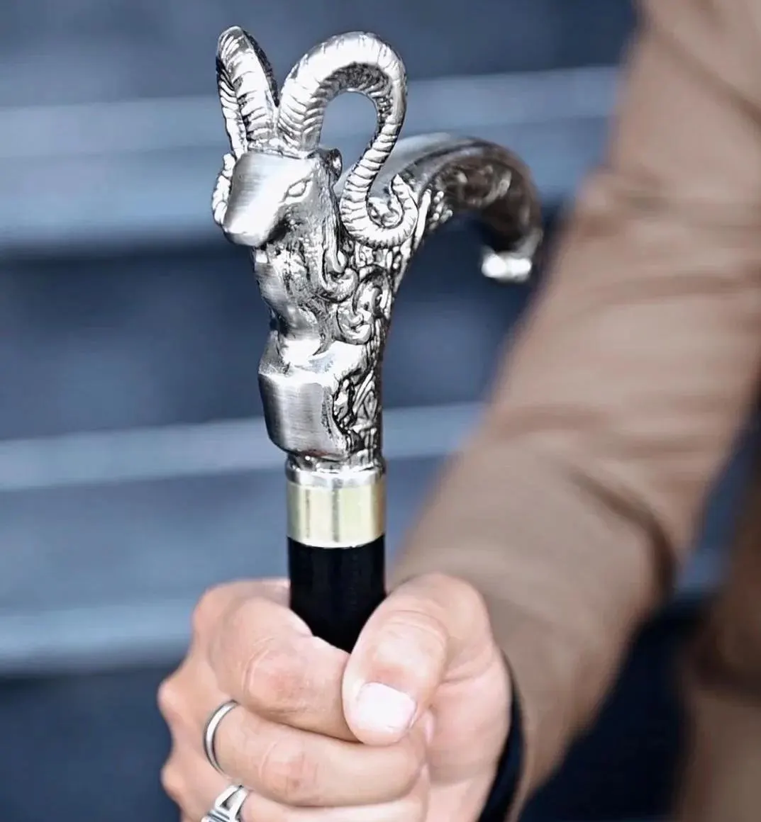 A walking stick featuring a silver-toned metal handle shaped like a ram head with detailed curved horns and ornate scrollwork. The handle is attached to a smooth black shaft with a metallic gold-coloured collar at the connection point.