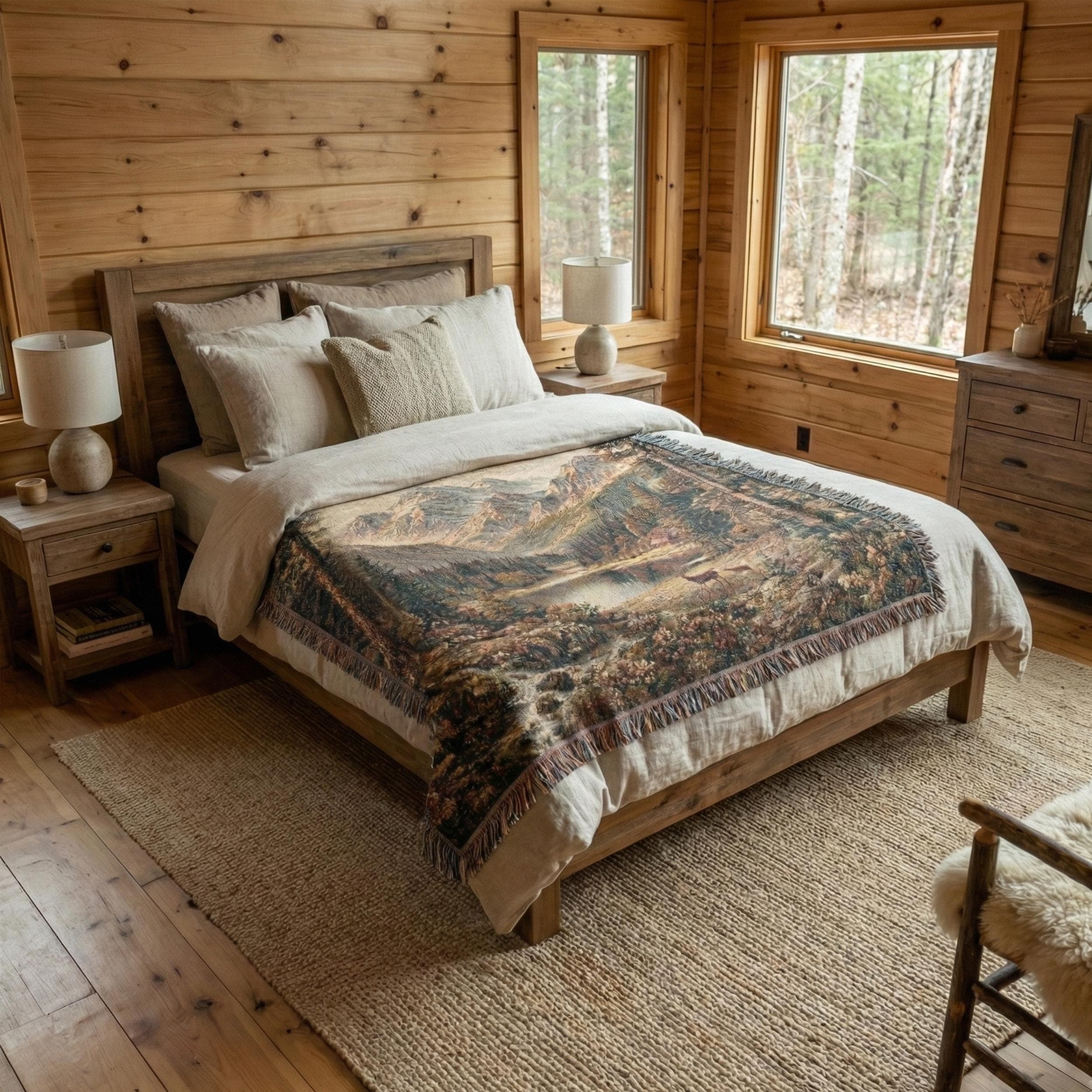 Woven tapestry blanket showing a vintage mountain landscape with an alpine lake in the center, evergreen forest on both sides, rocky stream in the foreground, and layered mountain peaks in the distance. Warm earthy tones and detailed woven texture with fringed edges. Works as tapestry wall art or woven wall hanging for cabin lodge decor, rustic home style, mountain house wall decor, and nature-inspired bedroom or living room accent. Great gift for outdoor lovers.