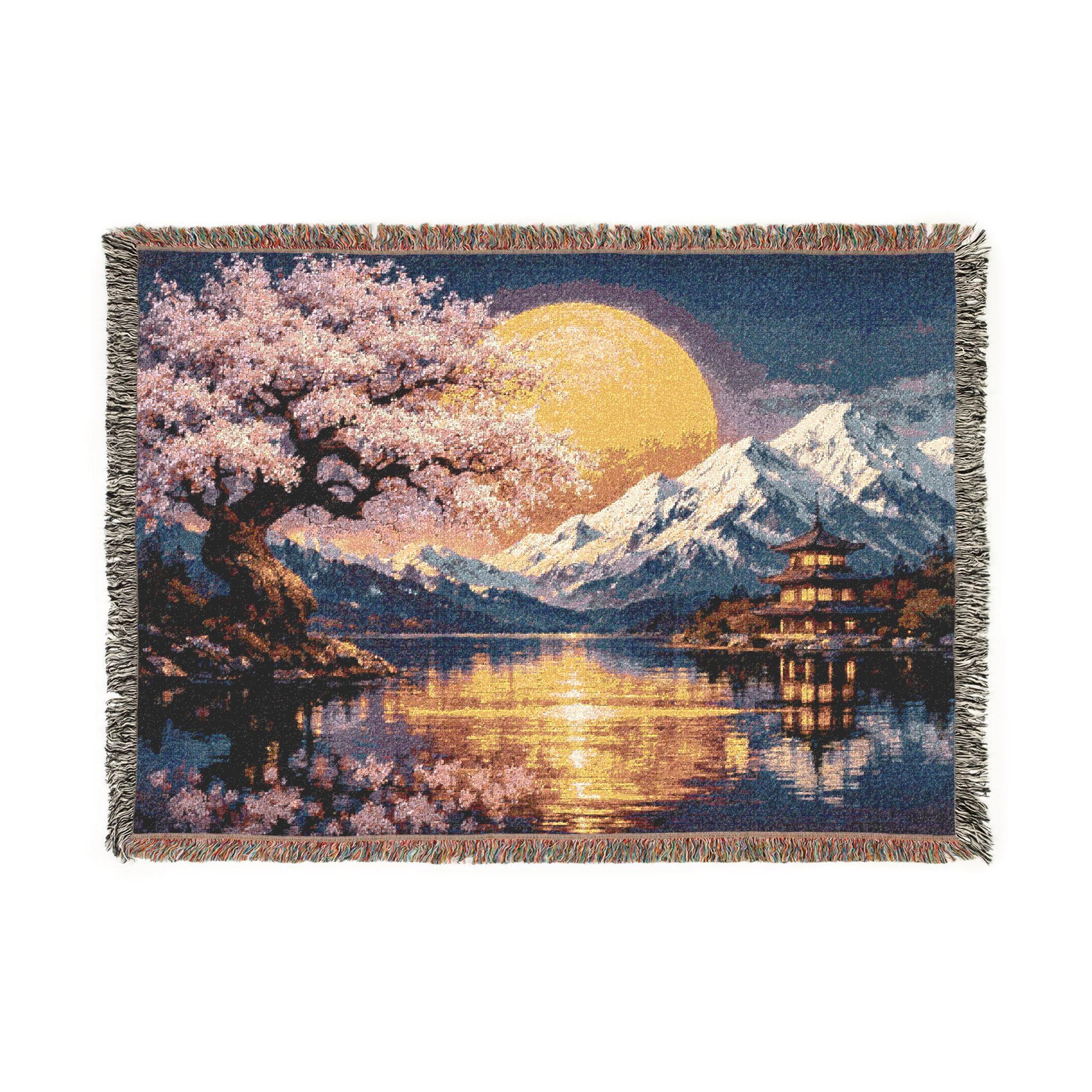 Woven tapestry blanket featuring a Japanese-inspired night landscape with pink cherry blossoms, a large golden moon, snowy mountains, and a pagoda beside a calm lake with shimmering reflections. Deep blue sky and warm light create a serene, zen mood. Fringed edges and detailed woven texture make it ideal as tapestry wall art, wall hanging, or cozy throw for bedroom, living room, reading nook, meditation space, or Asian-inspired home decor.