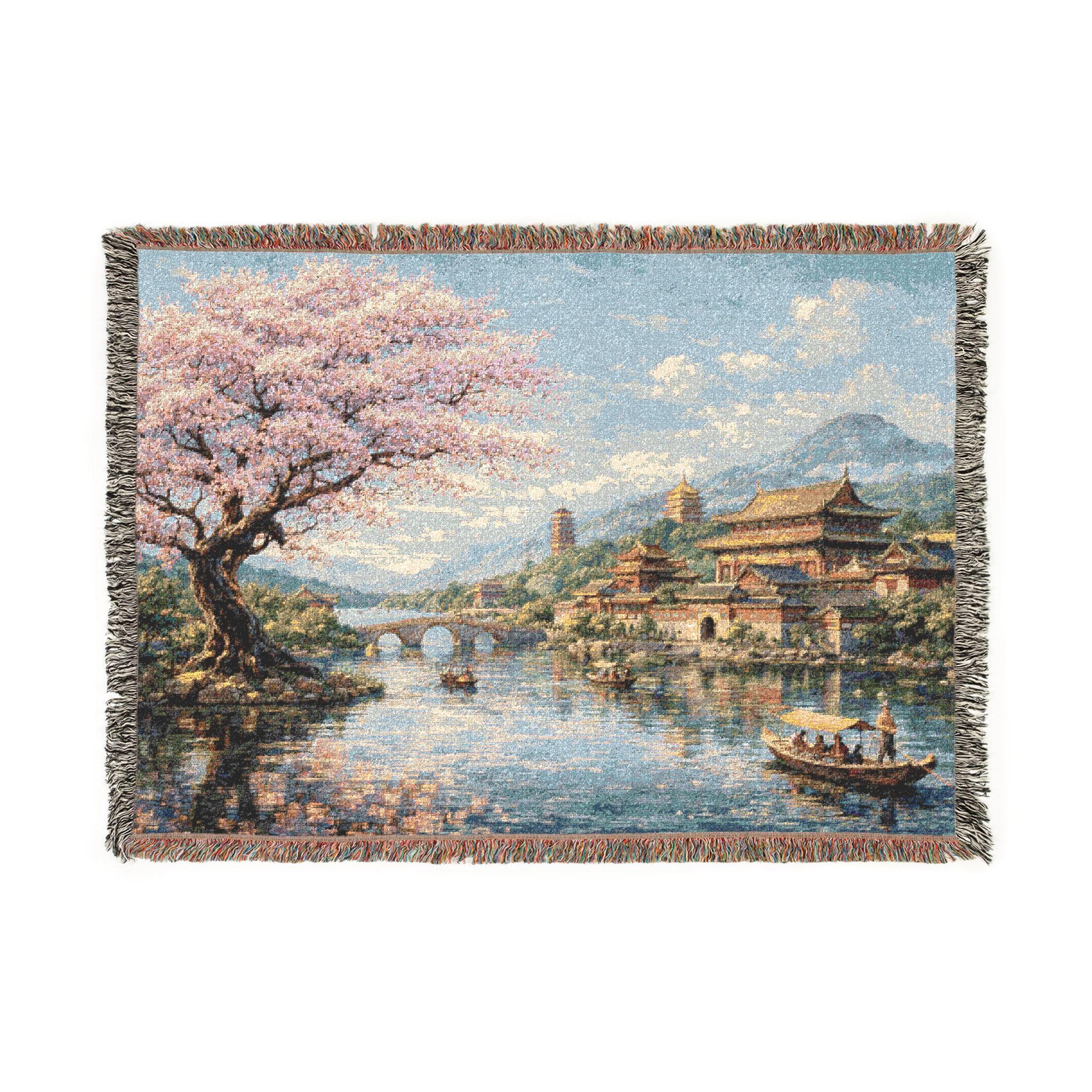 Woven tapestry wall hanging featuring a blooming cherry blossom tree beside a peaceful river with elegant Chinese palace architecture, pagoda style towers, an arched stone bridge, distant blue mountains, and small wooden boats drifting across reflective water under a bright sky. Vintage inspired Asian landscape tapestry with soft pink spring blossoms, serene garden scenery, and refined old world detail, ideal as a large wall tapestry, woven throw blanket, and gift for decor and travel art lovers