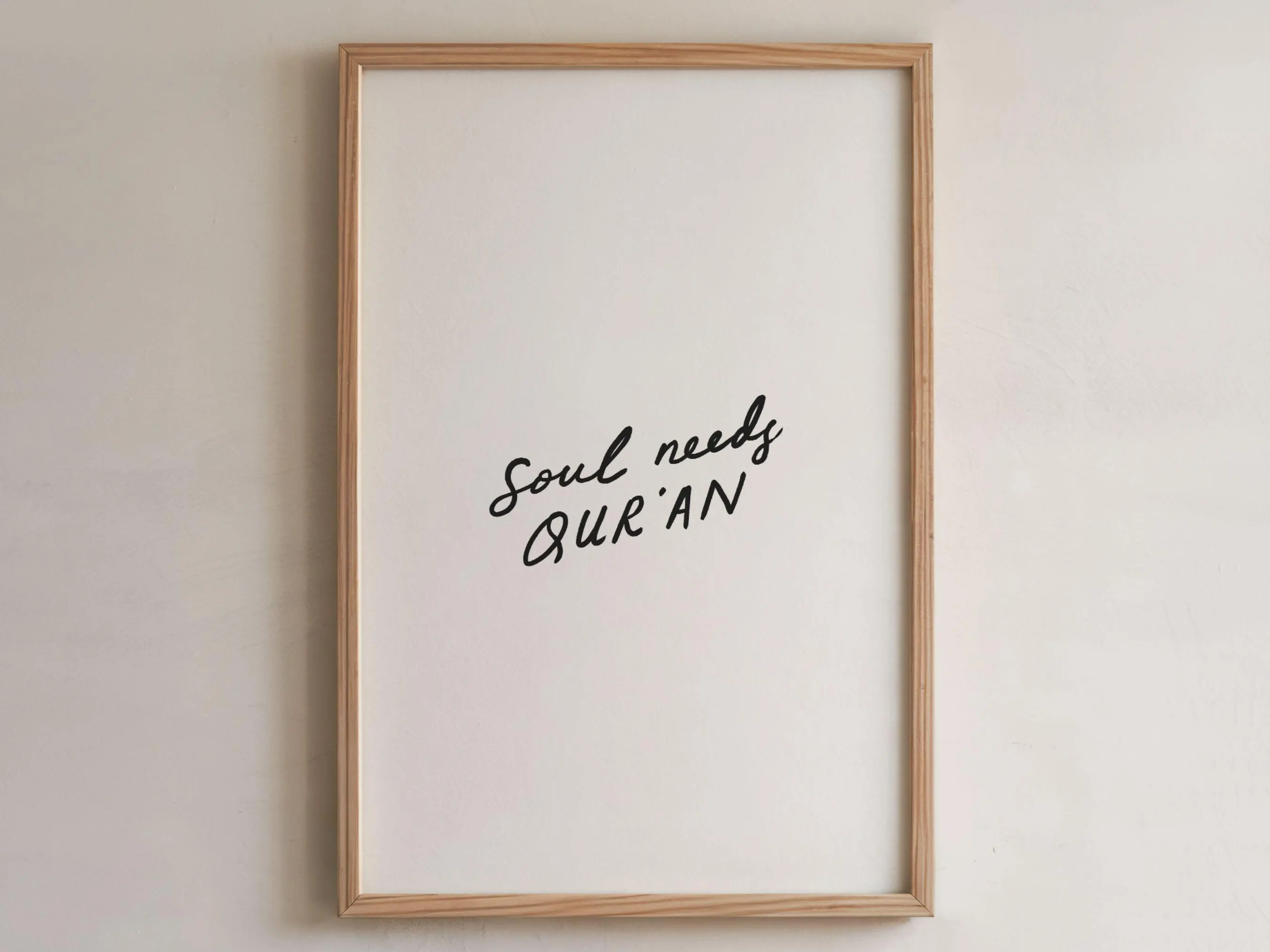 May include: A framed print featuring the words "Soul needs QUR'AN" in black handwritten script on a white background. The frame is made of light-coloured wood. The artwork is displayed on a neutral-coloured wall.