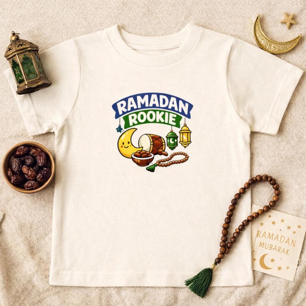 May include: Cream-colored t-shirt featuring a colorful graphic with the text "Ramadan Rookie". The design includes a smiling crescent moon, a drum, lanterns, and a string of prayer beads. The shirt is a casual wear item.