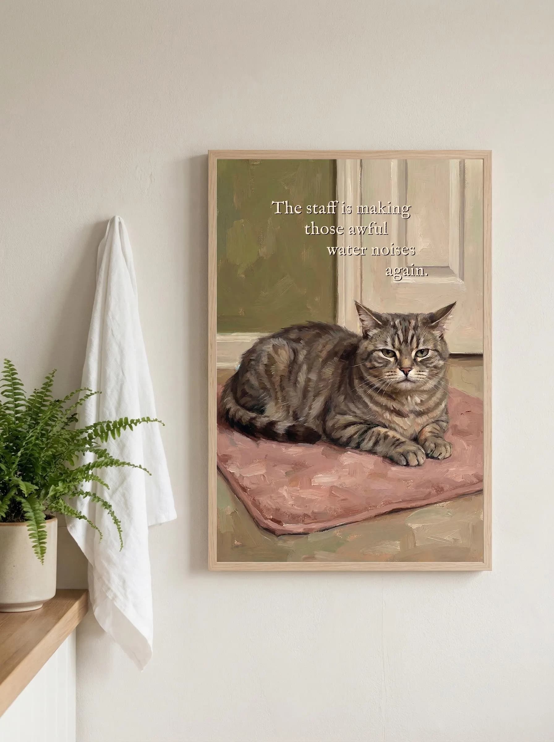 Cat humor wall art. Grumpy brown tabby cat with green eyes lying on a pink bathroom mat outside a closed white door, oil painting style, muted olive and pink palette. Quote: "The staff is making those awful water noises again." Funny bathroom signs style wall art.