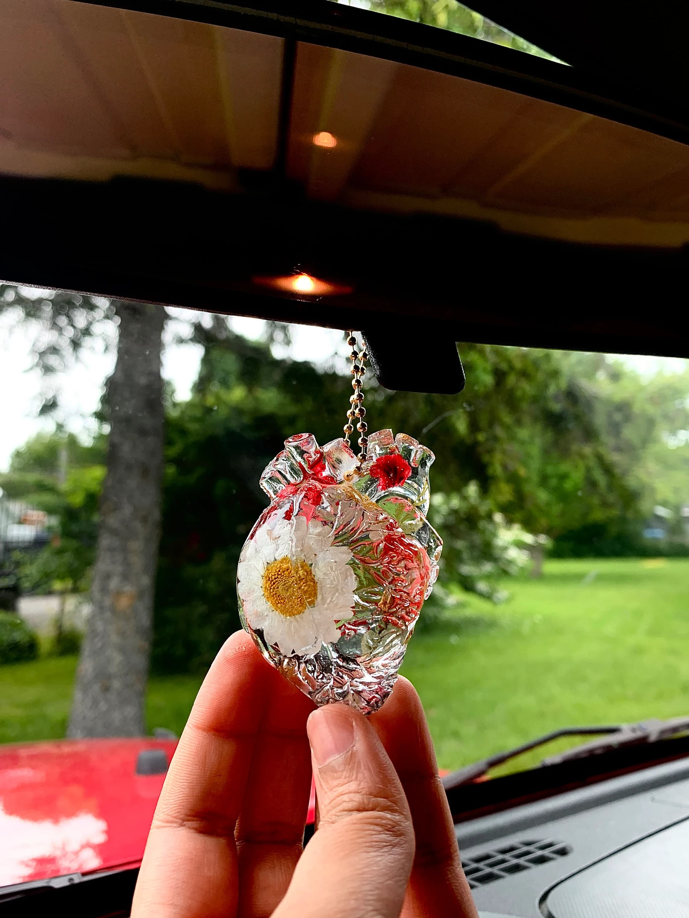 May include: A clear resin heart-shaped ornament with a white daisy and red flowers inside. The ornament is hanging from a silver chain.