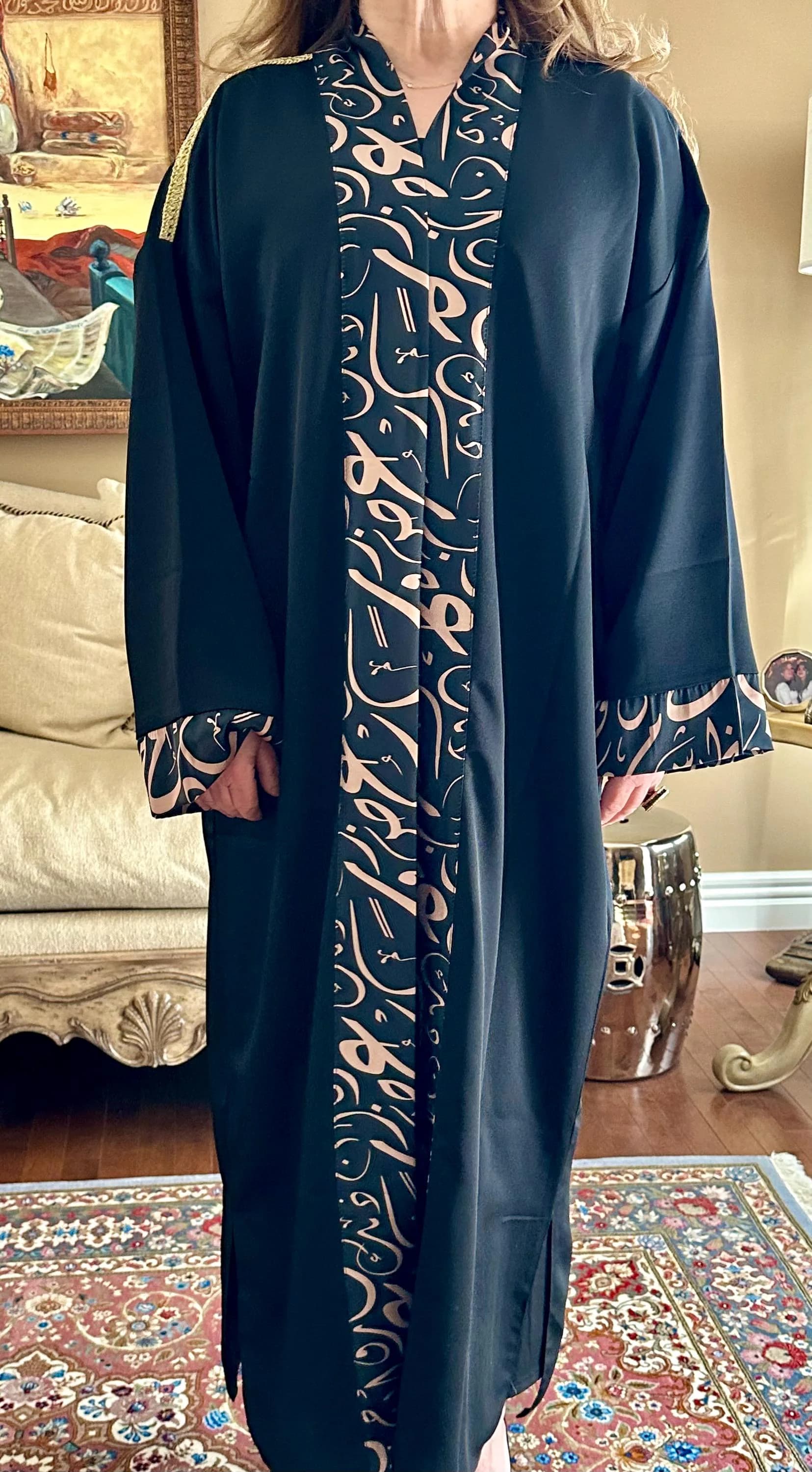 May include: Black abaya with a beige calligraphy design down the front and on the cuffs. The long-sleeved garment has a flowing silhouette and is suitable for modest wear. Gold trim accents the shoulders.